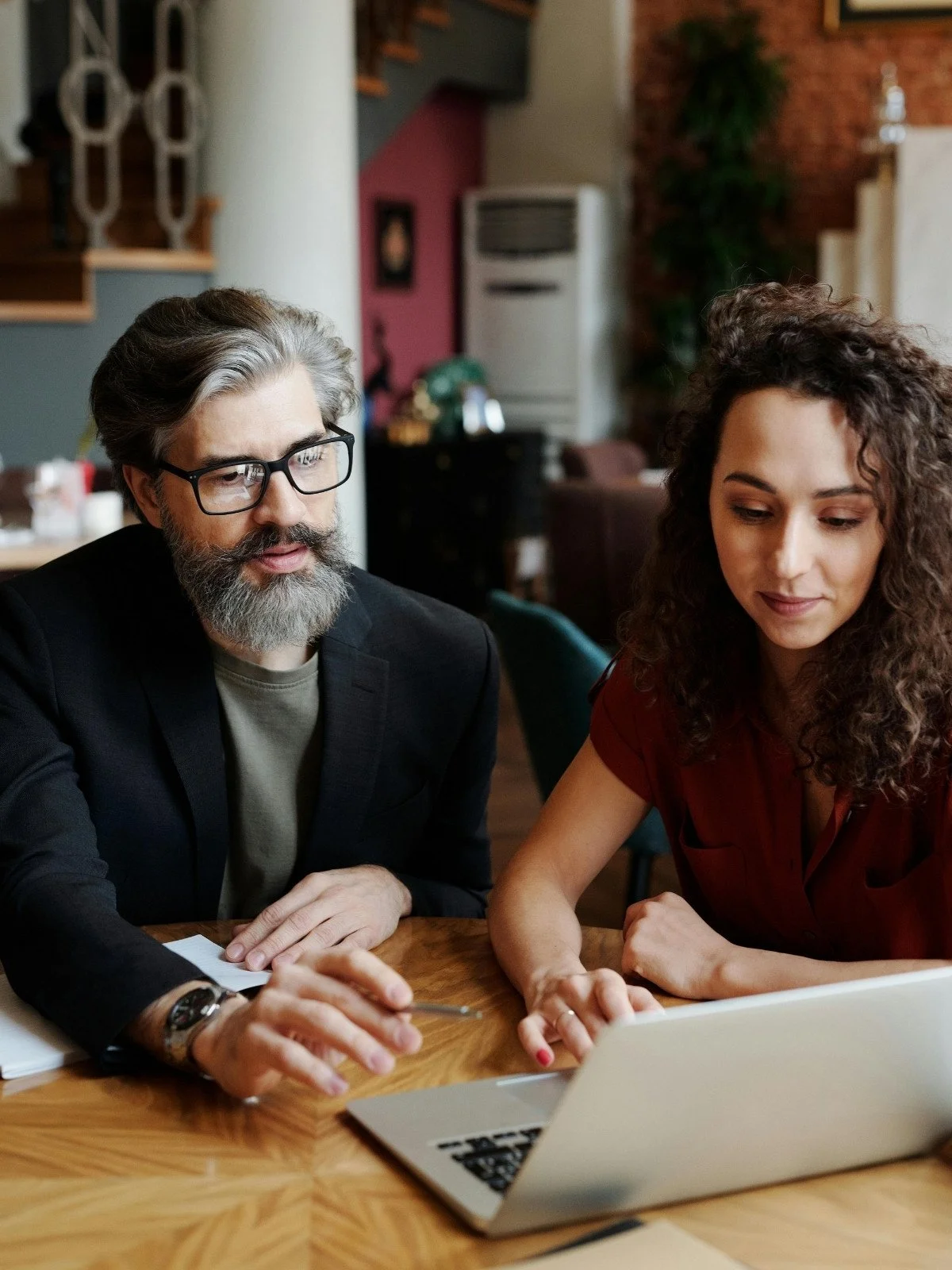 A middle-aged man with glasses and a beard, wearing a dark blazer and sitting at a wooden table, engages in a discussion with a young woman with curly hair wearing a red blouse, both focused on a laptop in front of them.