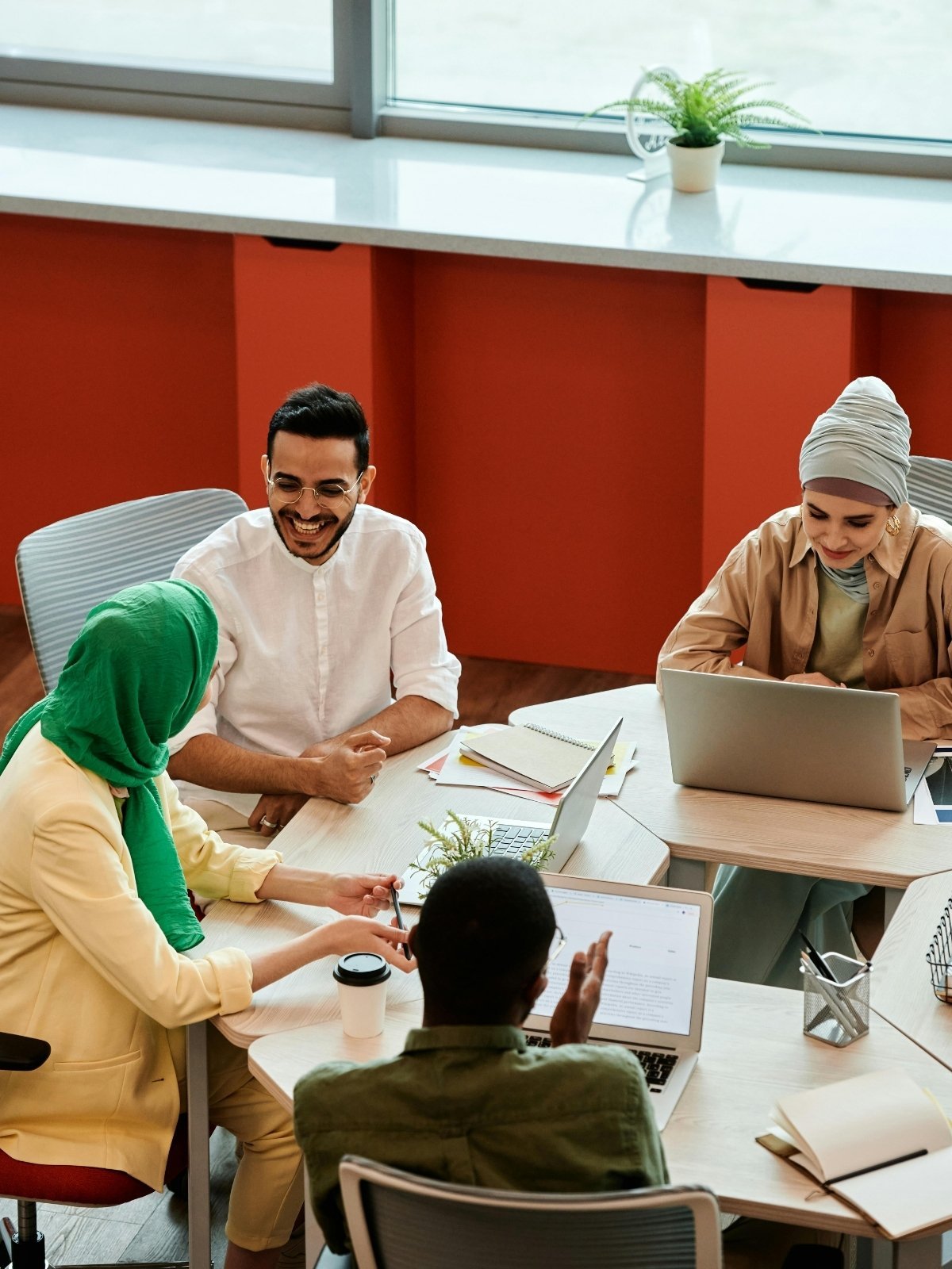 A diverse group of five people sitting around a table in a modern office, engaged in a discussion, with laptops, notebooks, and coffee cups visible.