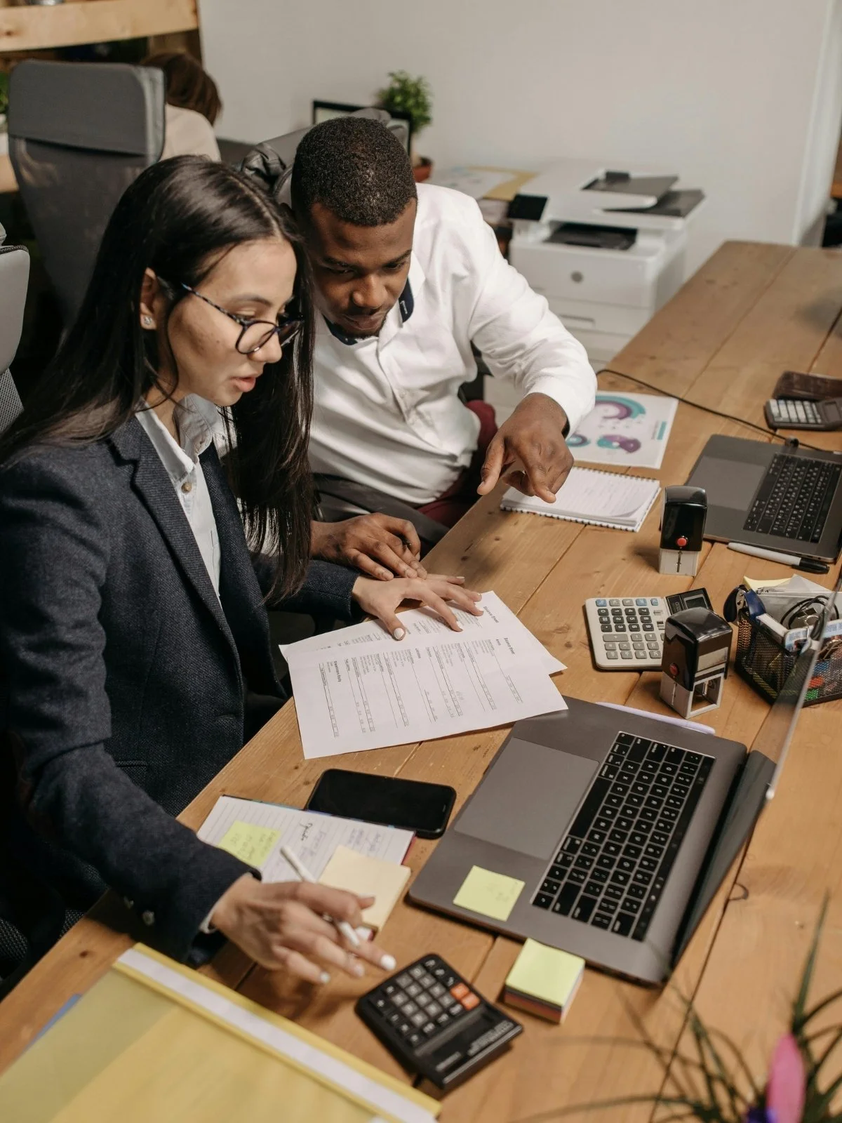 Two professionals reviewing documents at a cluttered desk with laptops, calculators, and office supplies.