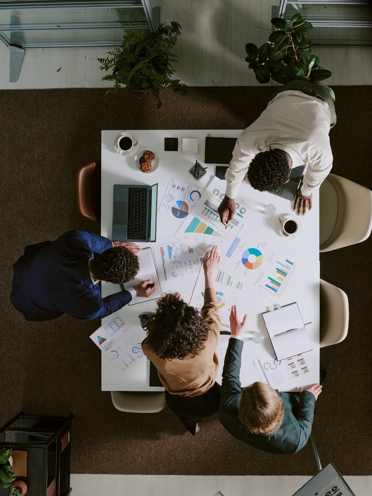 Women and men working together on business charts and graphs at a conference table, with laptops, coffee, and snacks.