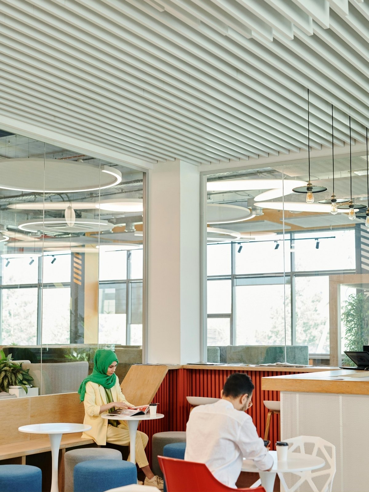 Modern cafe interior with large glass windows, contemporary lighting fixtures, and people sitting at tables, including a woman with a green hijab reading a magazine.