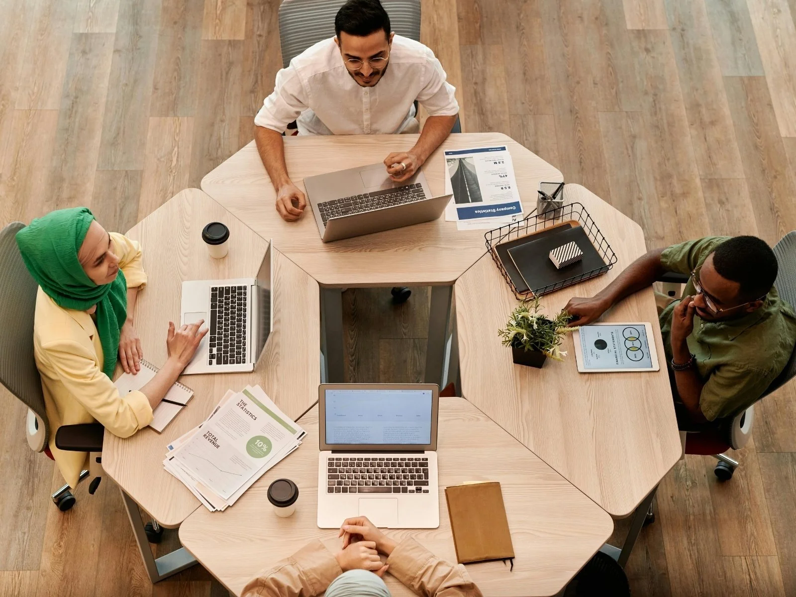 People sitting around a hexagon-shaped table in a meeting room with laptops, notebooks, documents, and a tablet.