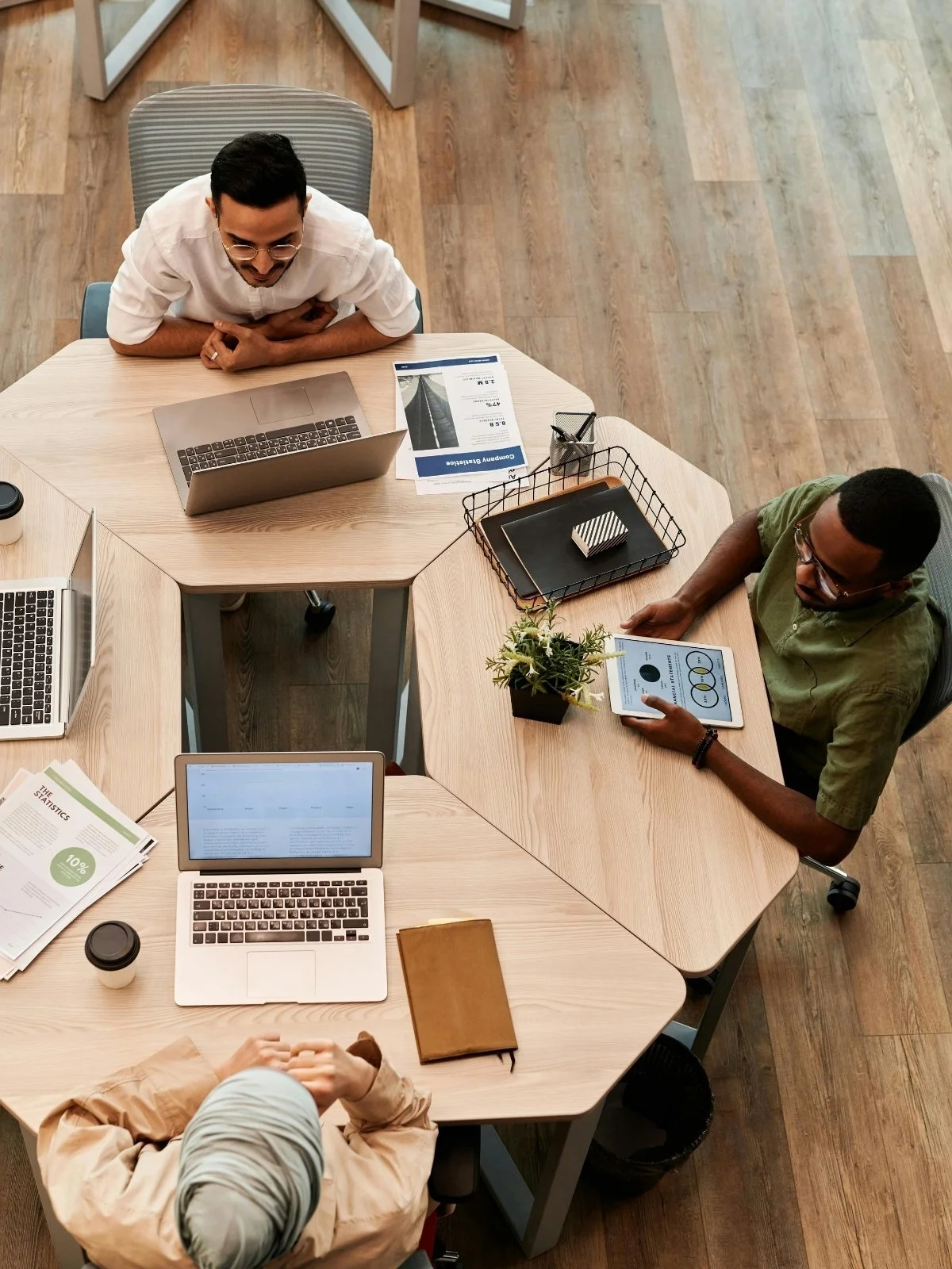 Top-down view of three people working at a wooden conference table with various laptops, tablets, papers, and office supplies.