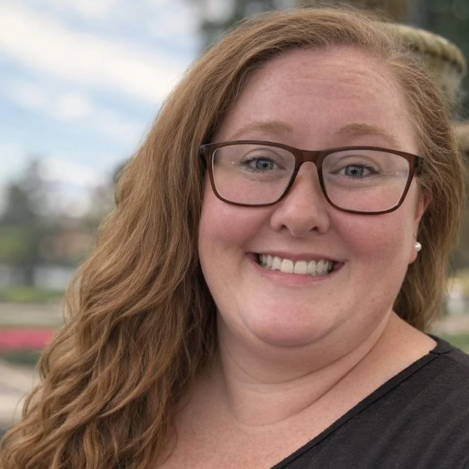 A woman with red hair, glasses, and earrings smiling outdoors on a cloudy day.