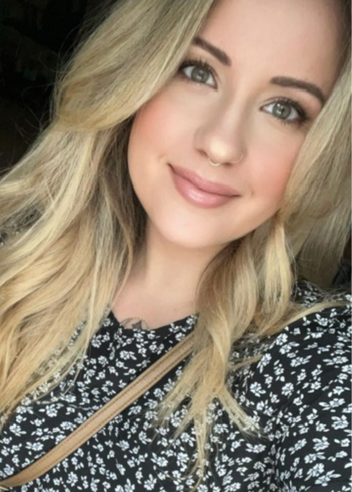 Close-up of a young woman with long blonde hair, gray eyes, and makeup, wearing a black dress with white floral pattern and a beige strap across her chest.