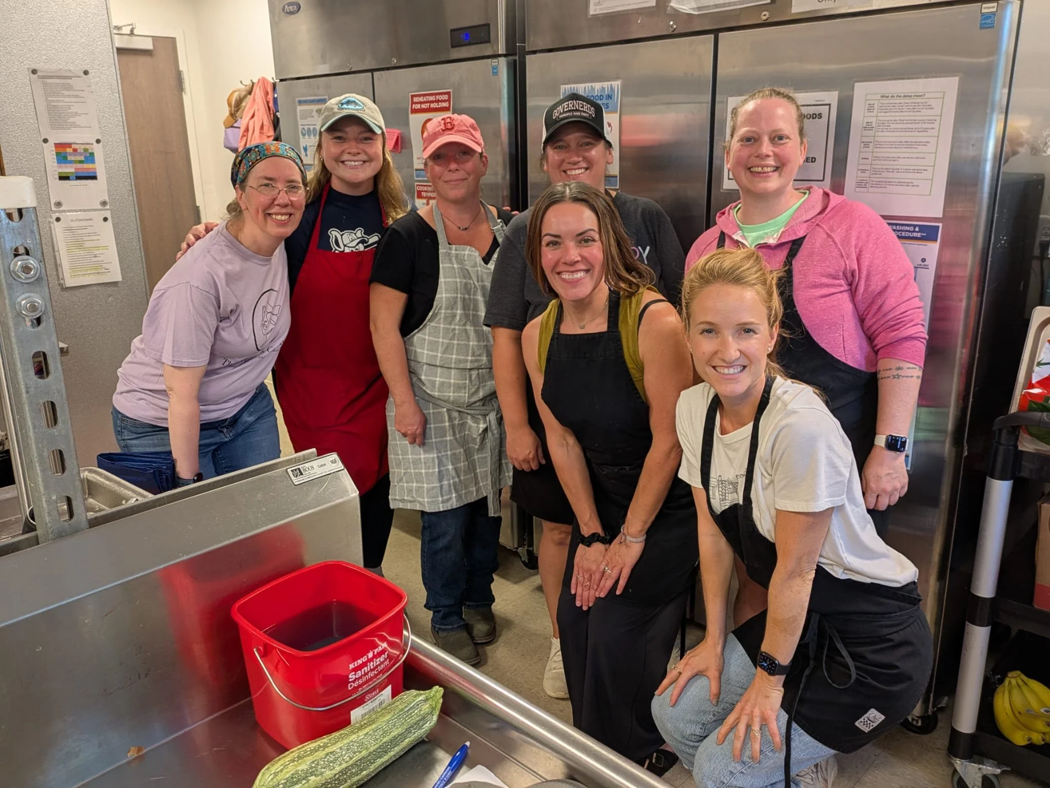 Group of seven women smiling in a kitchen setting, some wearing aprons and hats, with commercial refrigerator appliances in the background and a red sanitizer bucket in the foreground.