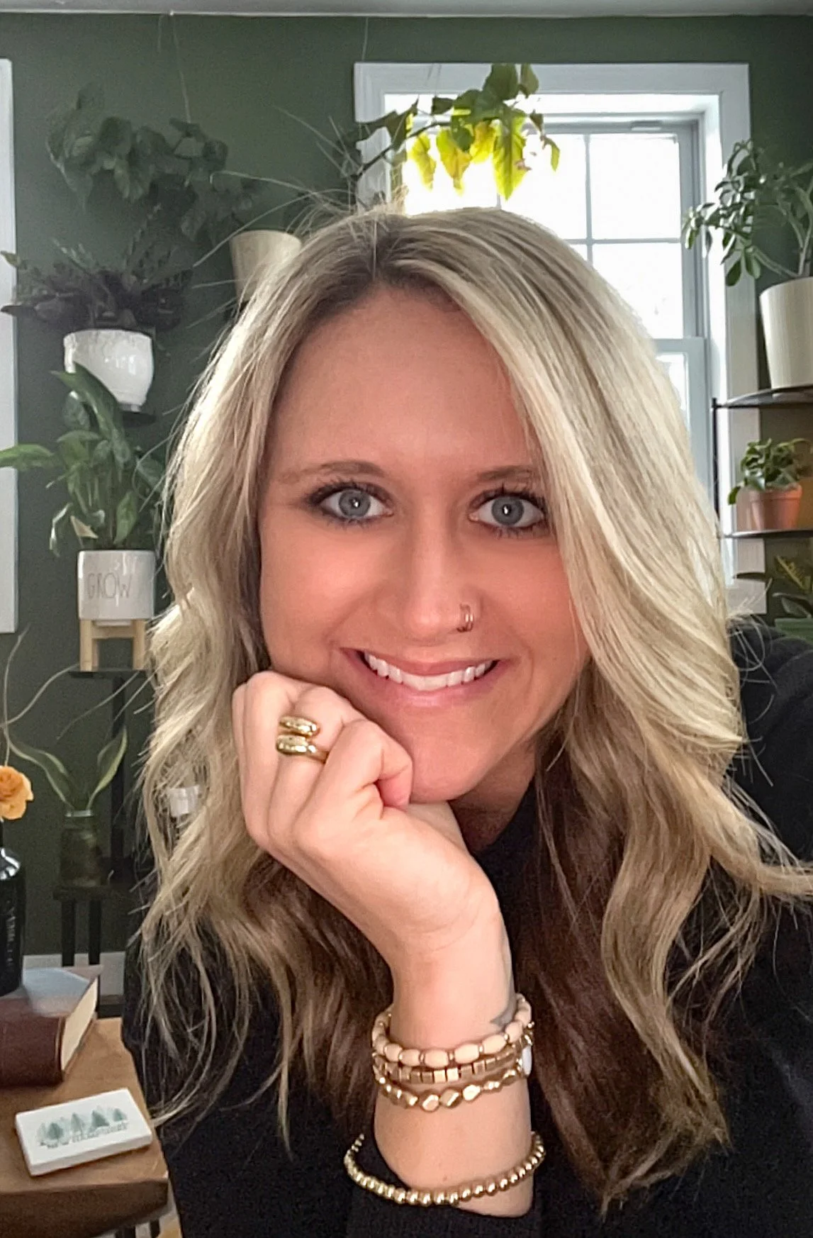 A young woman with long blonde hair, blue eyes, and a nose ring, smiling and resting her chin on her hand. She is wearing multiple rings, bracelets, and a black top. The background shows a window, houseplants, and a shelf with decorative items in a home or office setting.