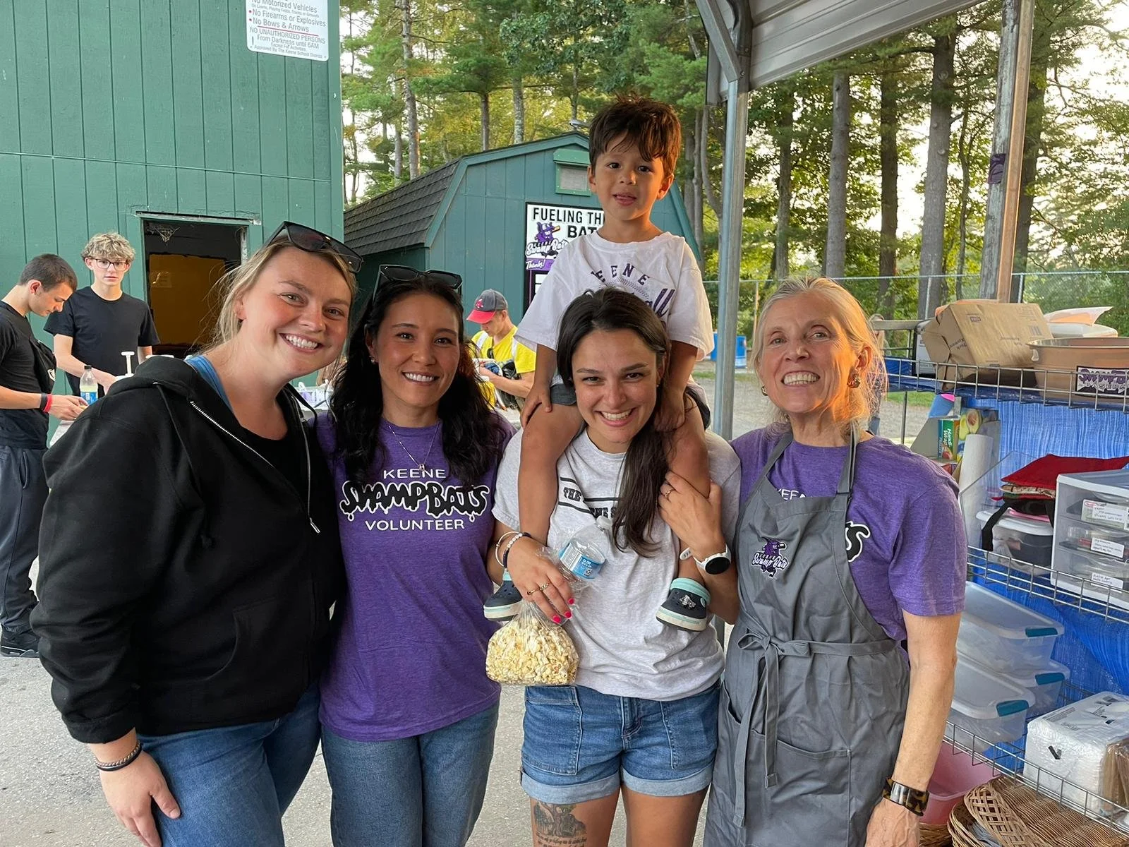 Five women and a young boy smiling at an outdoor event near a green building and food or supplies stand, with trees in the background.