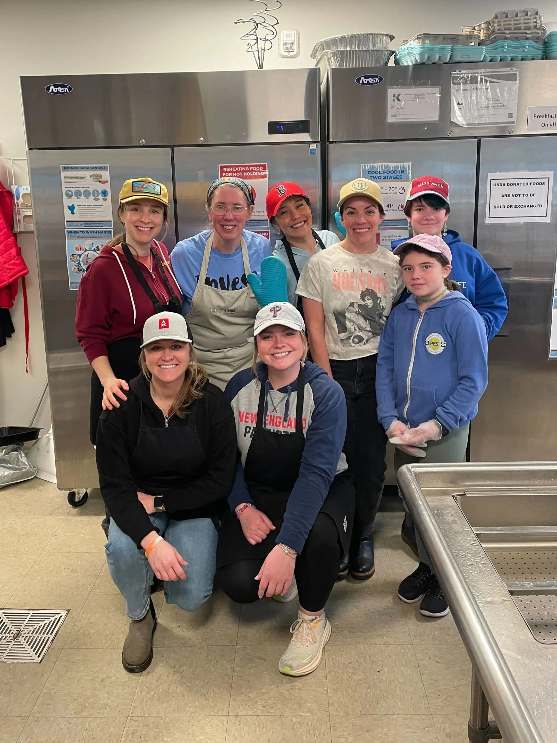 Group of eight women and girlsposing for a photo in a kitchen or food prep area, some wearing aprons and hats.
