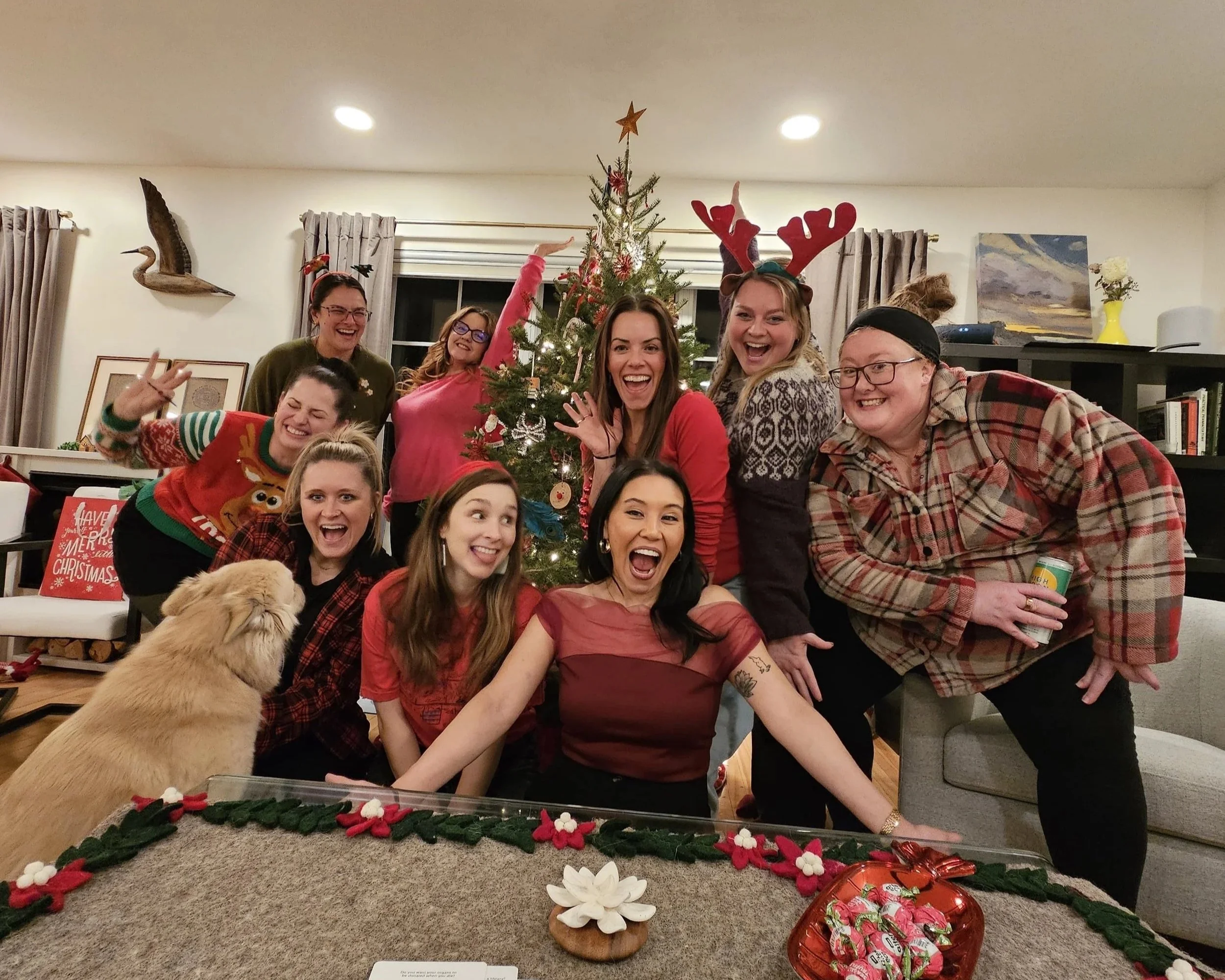 Group of women celebrating Christmas around a decorated Christmas tree, with some wearing holiday sweaters and reindeer antlers, smiling and posing enthusiastically in a cozy living room with holiday decorations.