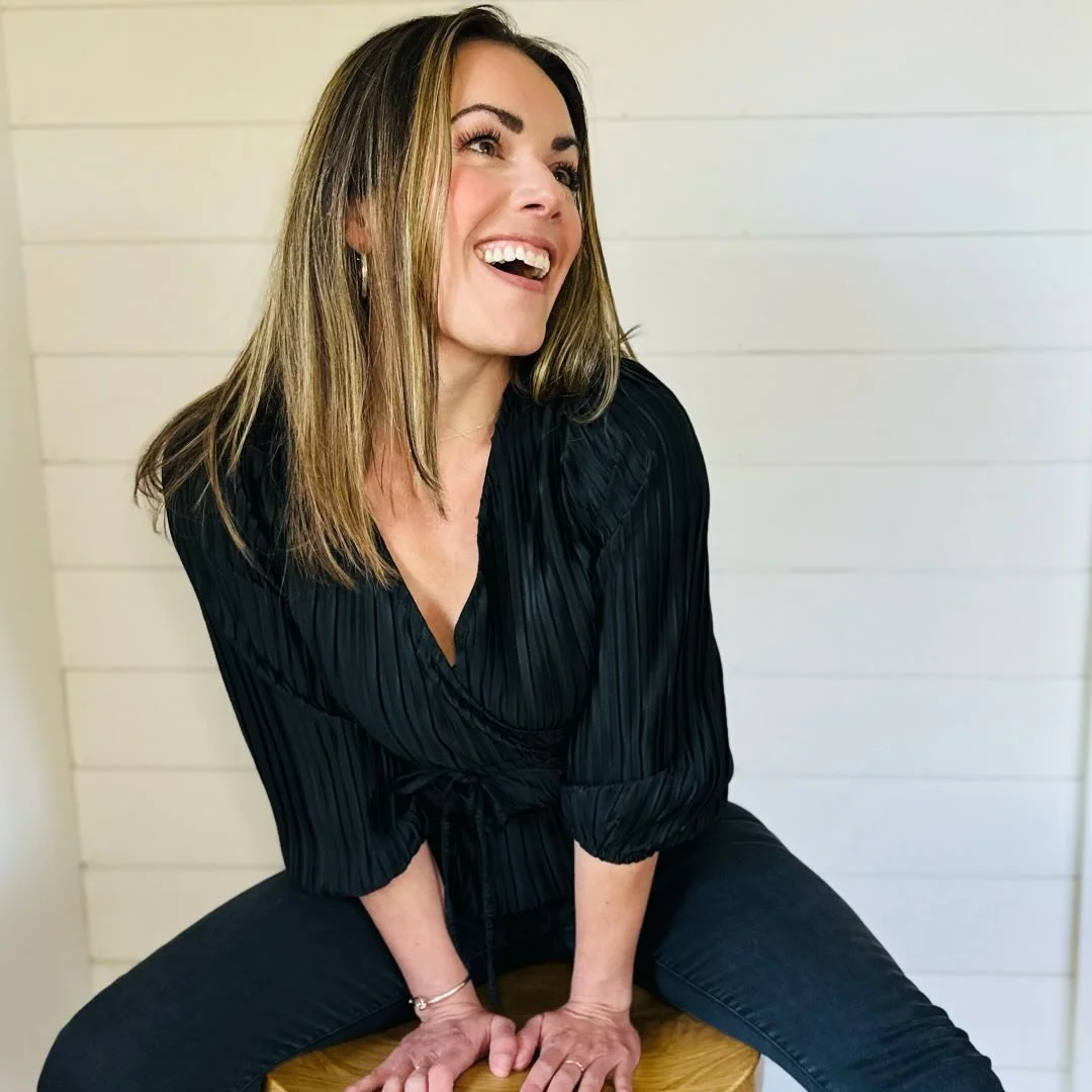 A woman laughs while sitting on a wooden stool, wearing a black pleated blouse and black pants, with a white shiplap wall background.