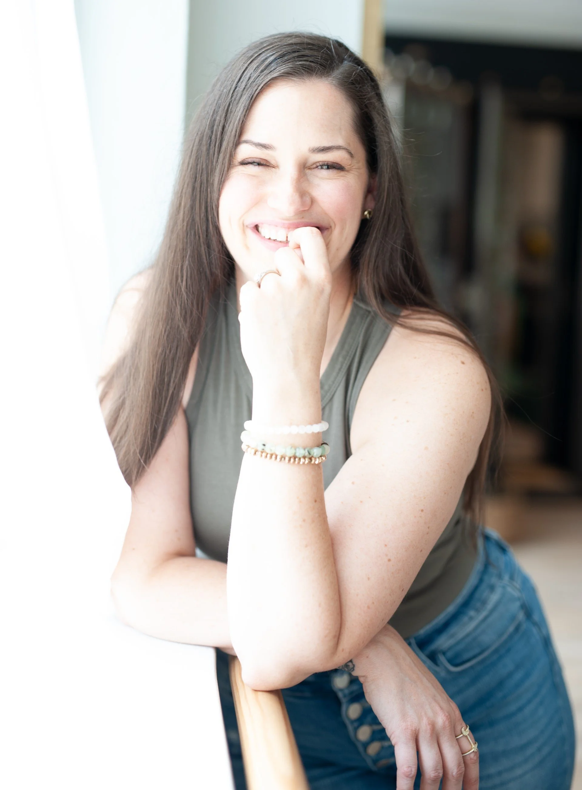 A woman with long brown hair, smiling, wearing a sleeveless top and denim jeans, sitting by a wooden chair with her hand near her chin.