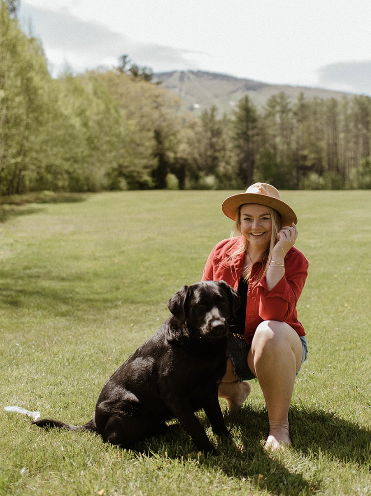 A woman with long blonde hair, wearing a wide-brimmed tan hat, red jacket, and denim shorts, kneels on green grass beside a large black dog in an open field. She is smiling, with a mountain and forested landscape in the background.