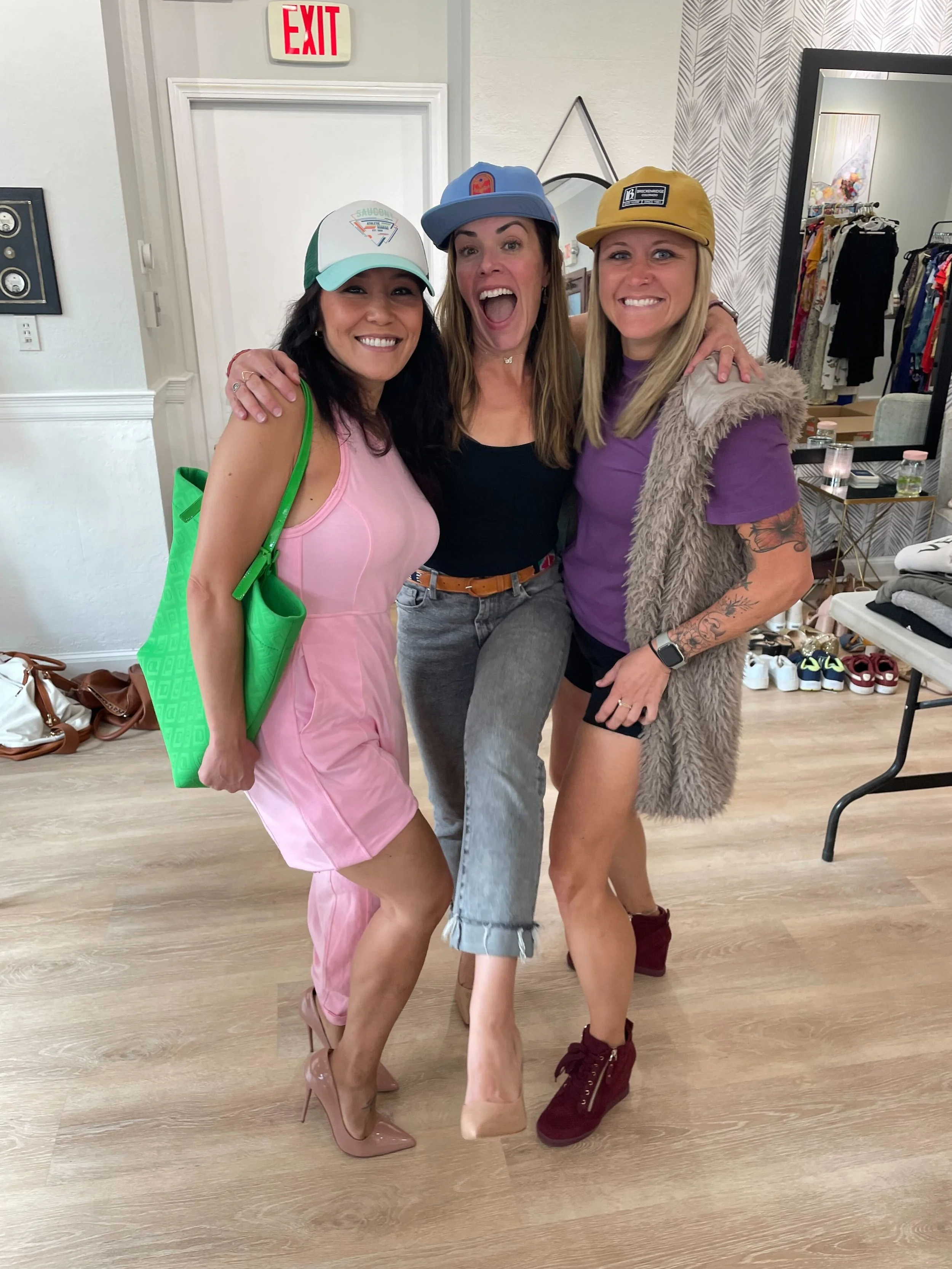 Three women standing close together, smiling, with arms around each other in a clothing store fitting room. The woman on the left wears a pink sleeveless dress and beige high heels, carrying a bright green bag. The woman in the middle wears a black top, gray jeans, and a blue cap. The woman on the right wears a purple T-shirt, black shorts, burgundy wedge shoes, a yellow cap, a furry vest, and has tattoos on her right arm.
