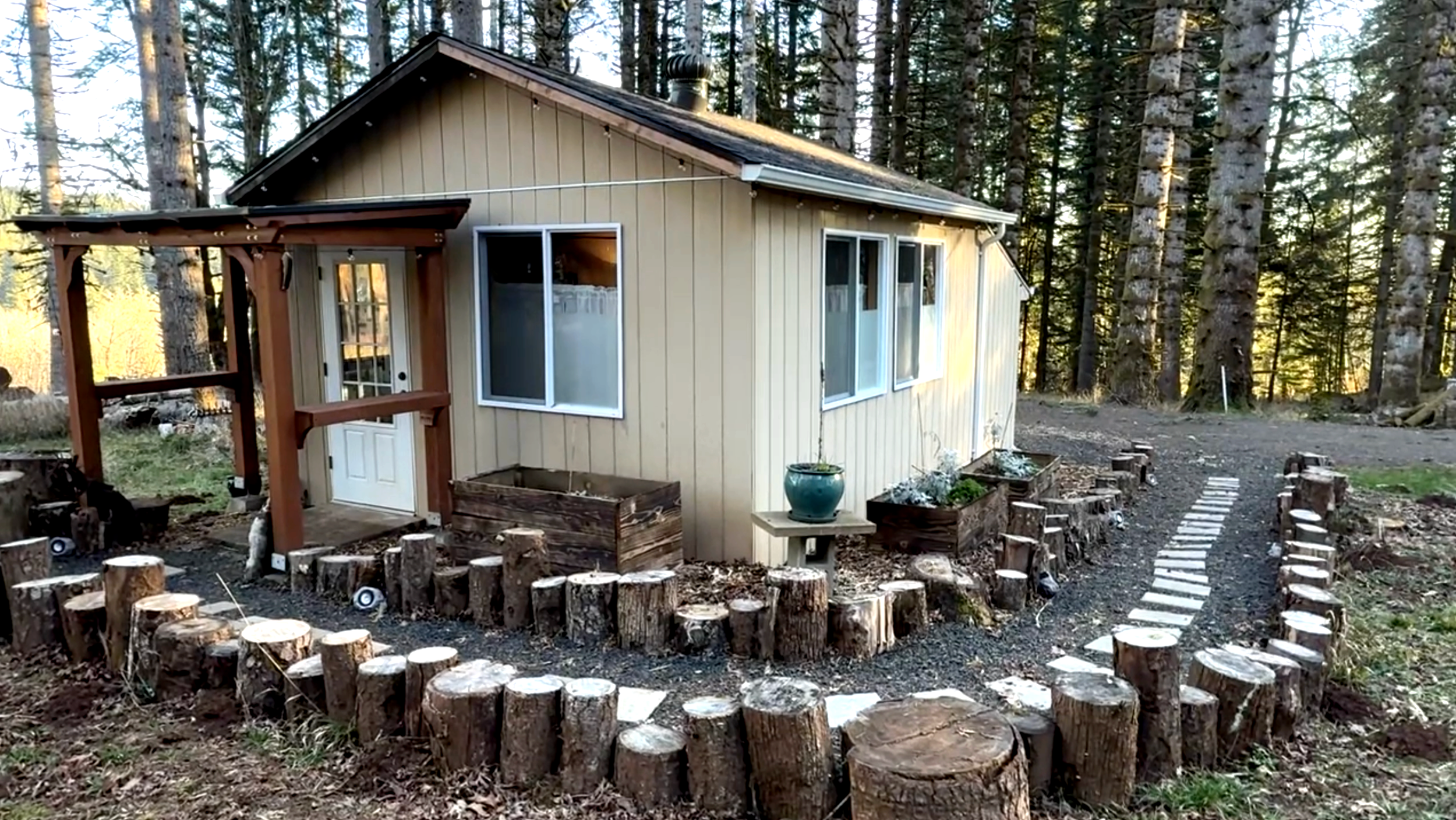 A small beige house with white trim, surrounded by a circular border made of cut logs, with a curved footpath leading away from it, in a wooded area.