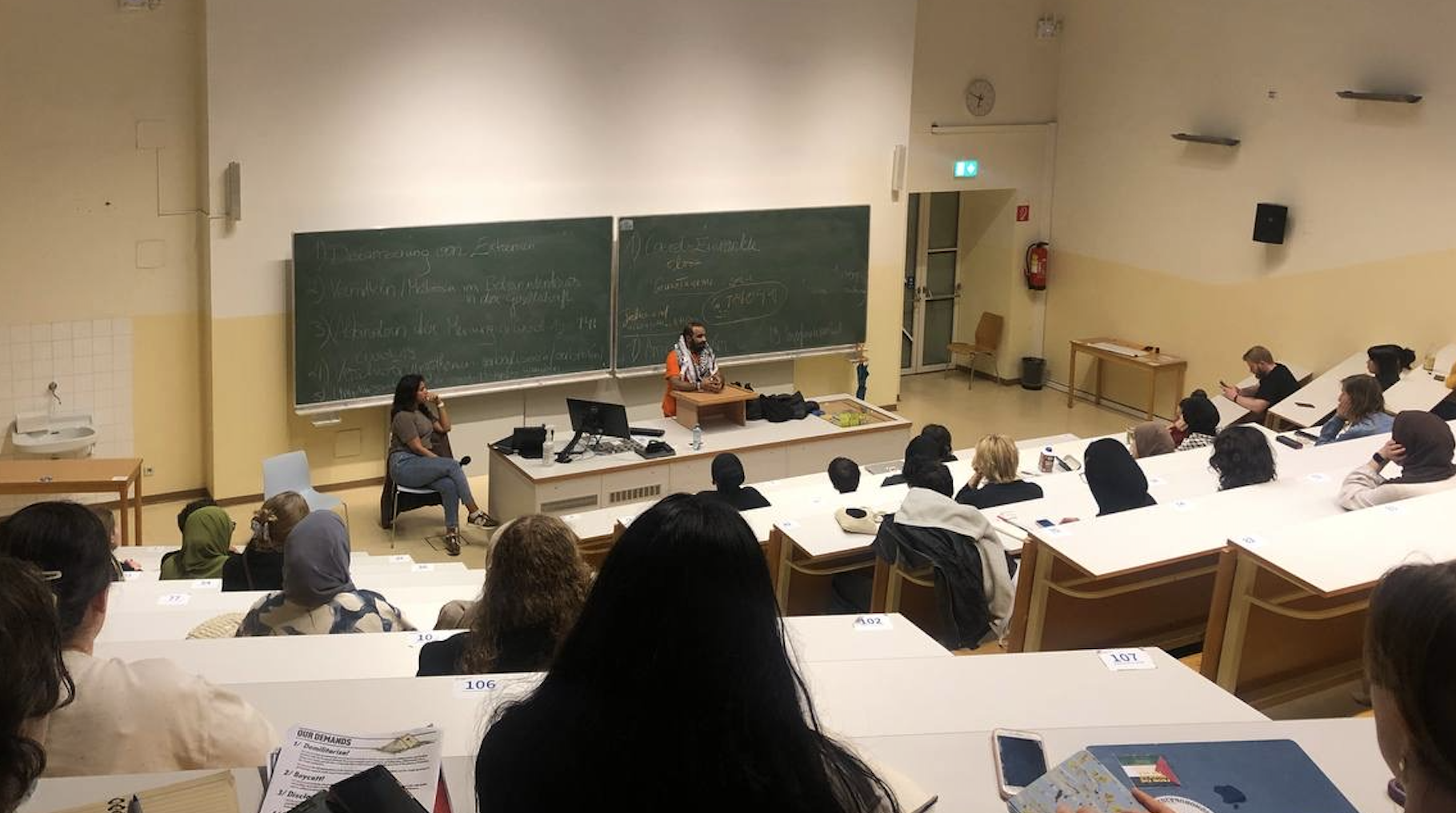 Universitätsvorlesung mit einem Lehrer an der Tafel, Studenten im Hörsaal, einige nehmen Notizen, ein Student hält ein Mikrofon, mehrere tragen Kopfbedeckungen, einige verwenden Handys, in der Ecke Feuerlöscher, Wanduhr an der Wand.