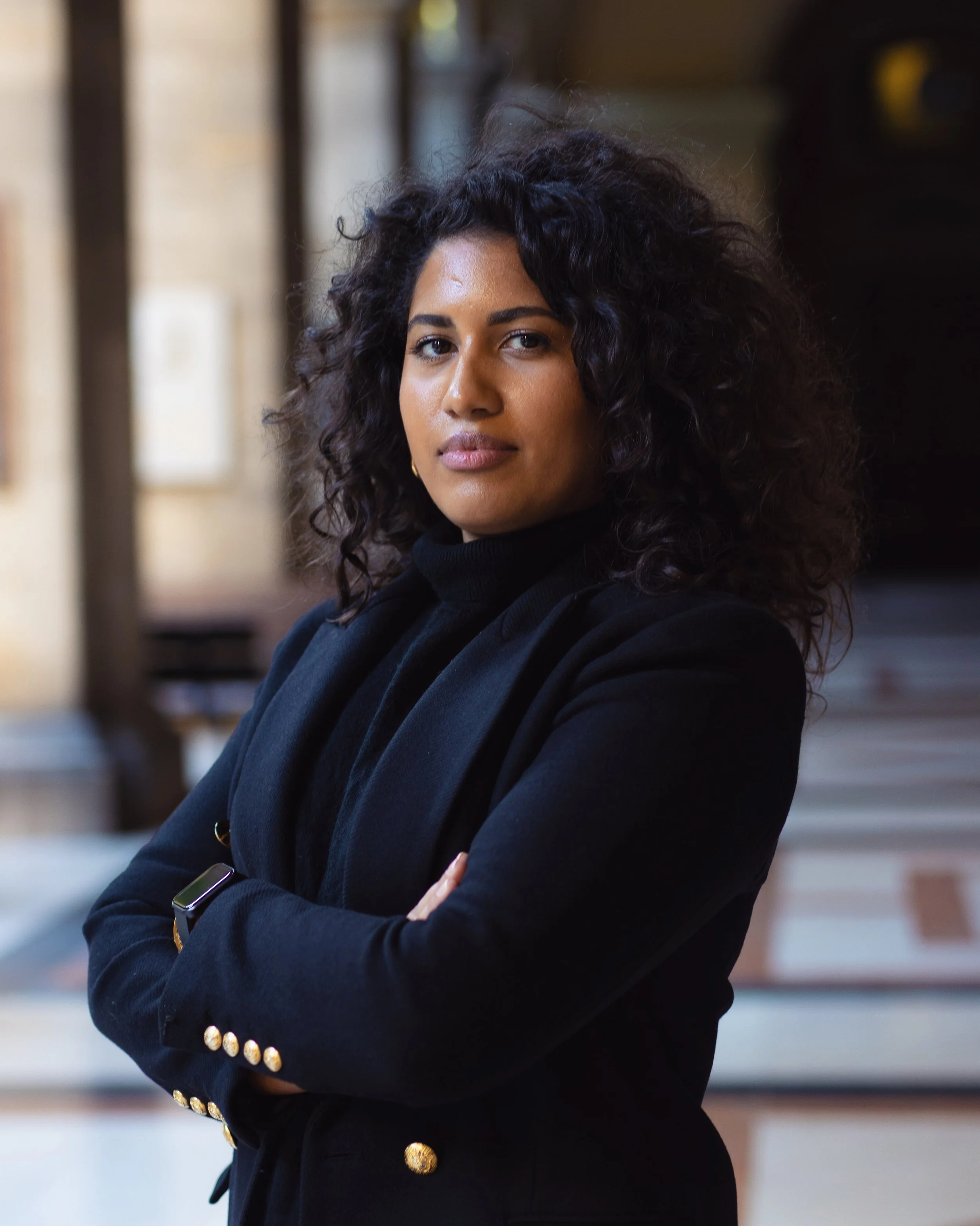 Selbstbewusste Frau mit lockigem schwarzen Haar, dunkler Kleidung mit goldenen Details, die Arme verschränkt in einem städtischen Umfeld