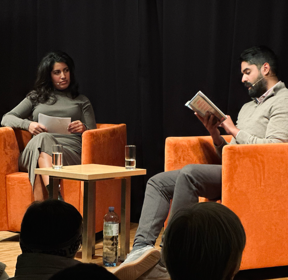 Zwei Menschen in einem Gespräch auf einer Bühne, eine Frau und ein Mann, beide tragen Headsets, im Hintergrund dunkle Vorhänge, vor ihnen auf dem Boden sitzen Menschen.