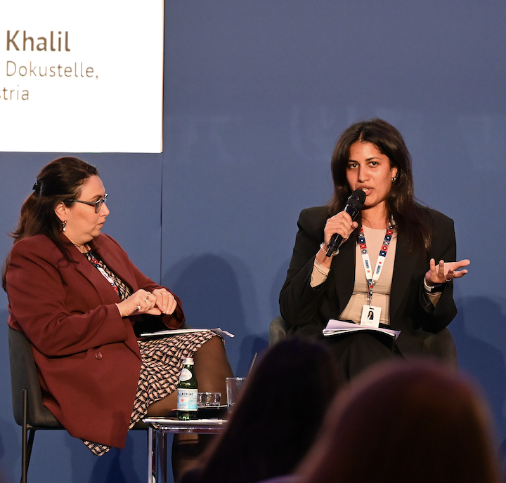 Zwei Frauen im Gespräch bei einer Konferenz, eine hält ein Mikrofon, die andere sitzt mit Notizen. Es ist ein Bühnen- oder Panel-Diskussion. Der Hintergrund ist blau, und auf einer Leinwand ist ein Teil eines Namens und einer Organisation sichtbar.