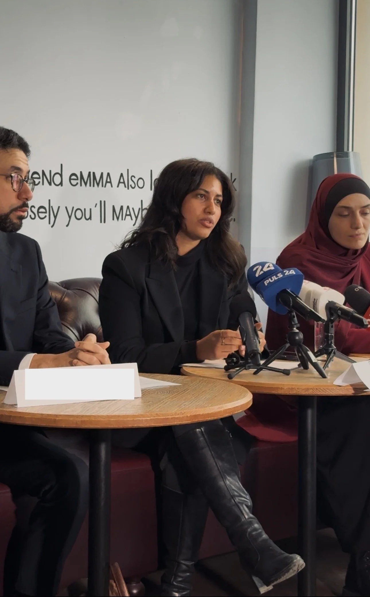 Drei Personen sitzen an einem runden Tisch bei einer Pressekonferenz, die Frau in der Mitte spricht, Mikrofone sind vor ihnen. Im Hintergrund ist ein Text, der teilweise sichtbar ist.