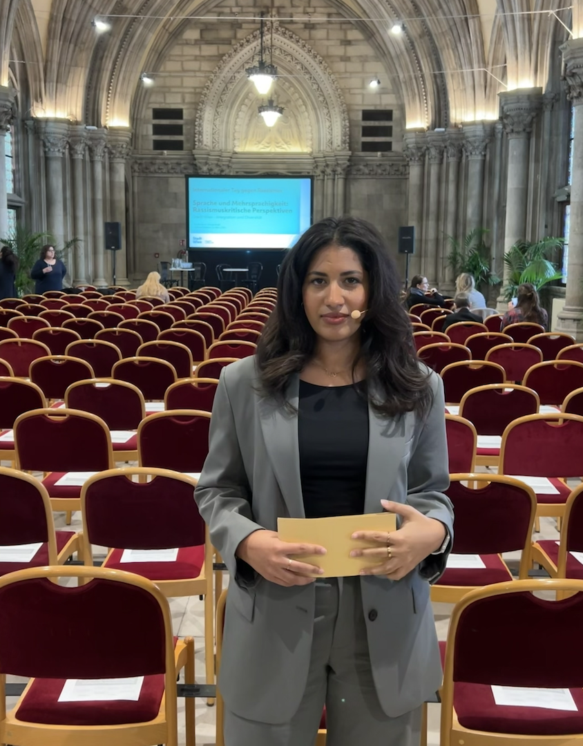 Eine Frau in einem grauen Anzug steht in einem großen, prunkvollen Saal mit roten Stuhllehnen, vor einer Leinwand, auf der Präsentationsfolien zu sehen sind. Es gibt mehrere Personen im Hintergrund, die an der Veranstaltung teilnehmen, und die Architektur ist architektonisch beeindruckend.