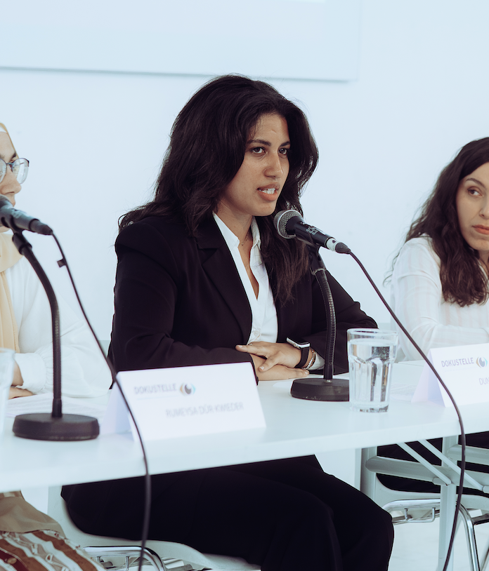 Eine Frau spricht bei einer Konferenz, sitzt an einem Tisch mit einem Mikrofon, Wasser und Namekarte.