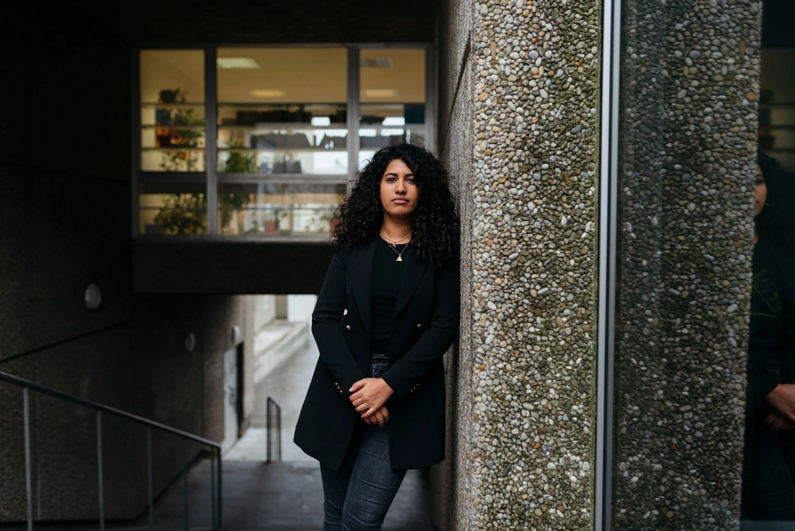 Junge Frau mit dunklen, lockigen Haaren trägt einen schwarzen Blazer und steht an einer Ecke eines Gebäudes mit einer Wand aus Kieselsteinen, im Hintergrund sind Fenster und eine Treppe sichtbar.