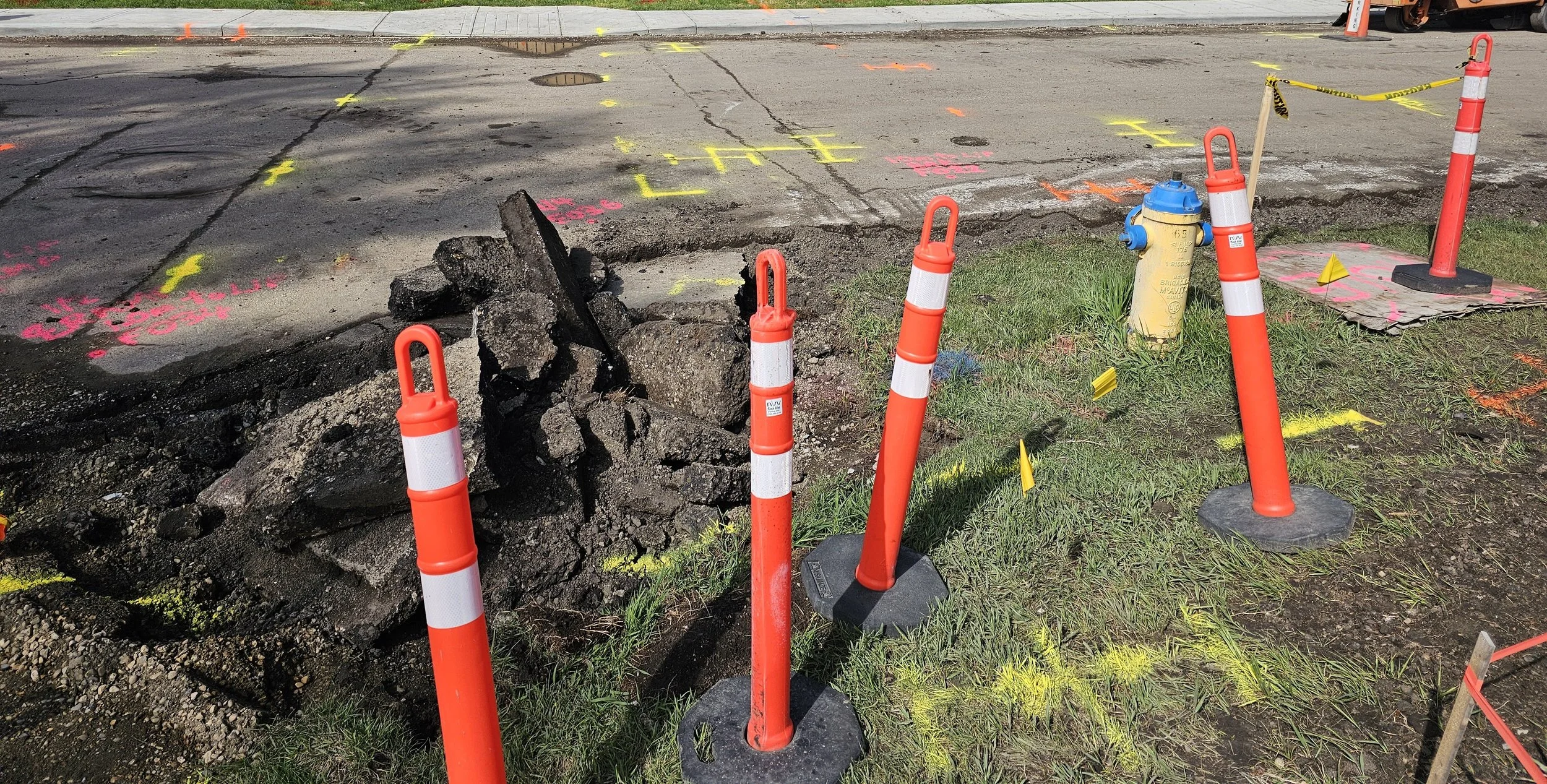 Sidewalk with an excavation surrounded by orange safety cones, yellow fire hydrant, and various paint markings in pink and yellow on the pavement identifying underground utilities and facilities.