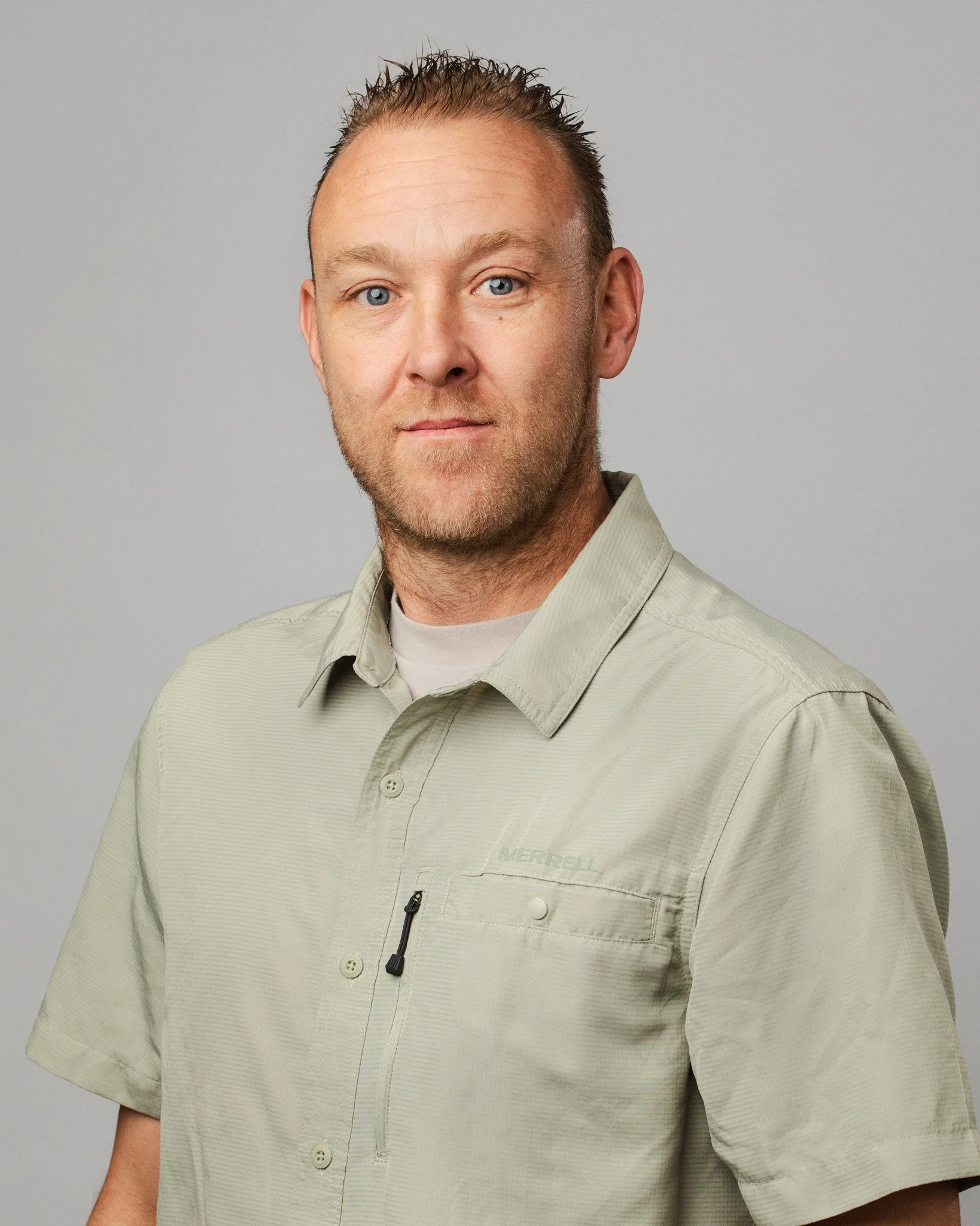 A man with short, spiked hair and a beard, wearing a light green button-up shirt, posing against a plain gray background.