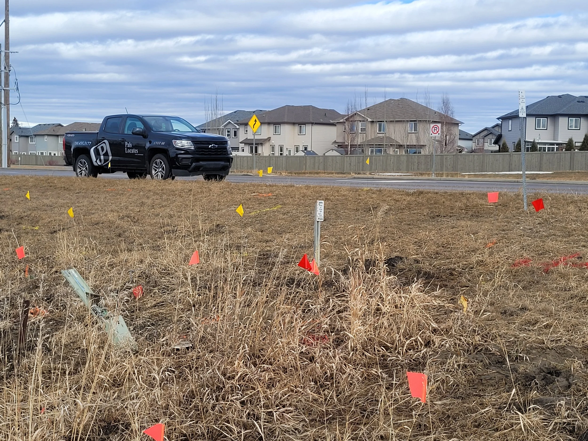 A patch of dry grass with small red and yellow flags marking underground utilities and facilities, indicating a construction site where ground disturbance will occur.  In the background a black truck parked on a residential street under a cloudy sky.