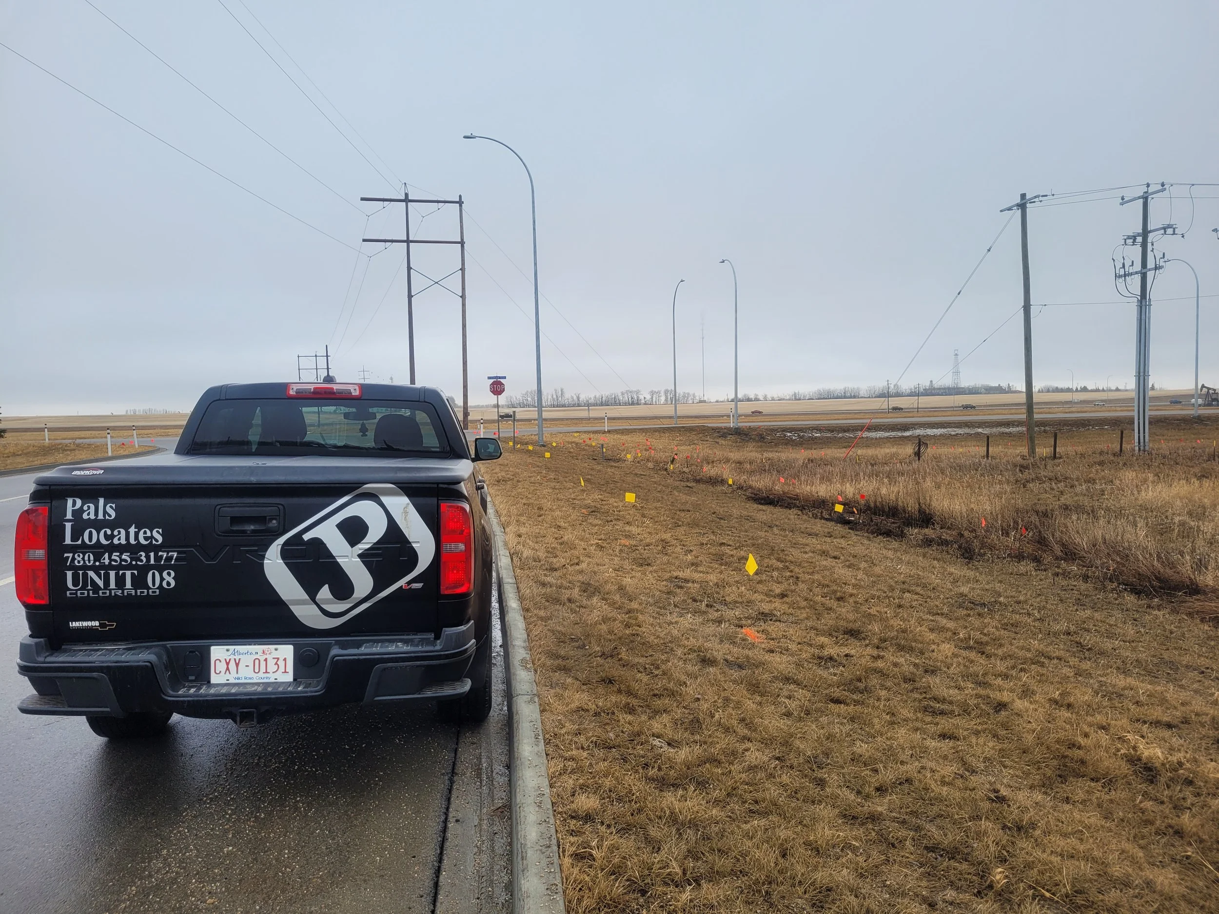 Black pickup truck with advertisement for Pals Locates and contact info parked on the side of a wet road in a rural area with power lines and yellow and red-marked stakes and flags in the grass.