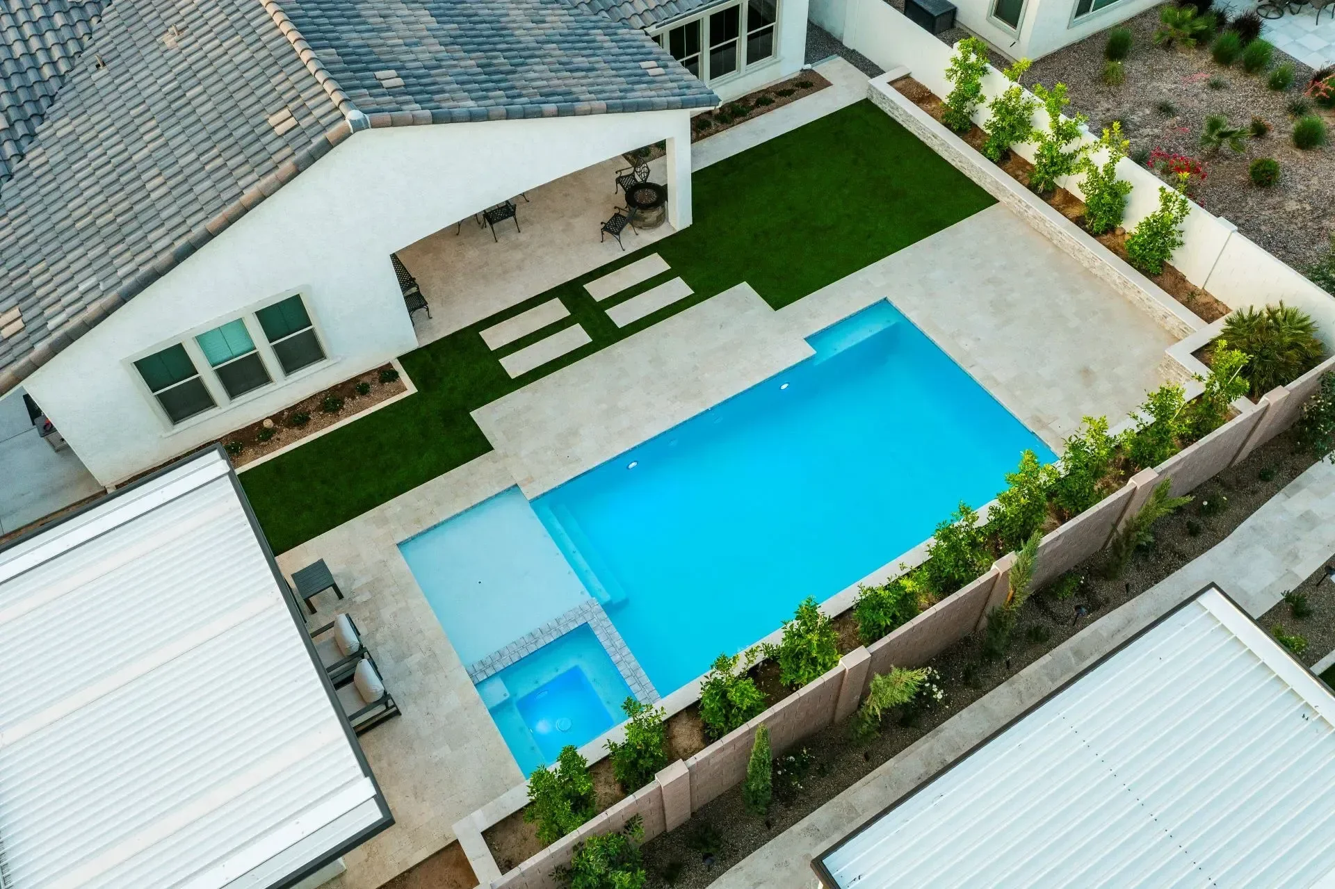 Aerial view of a backyard with a swimming pool, hot tub, patio, green lawn, and garden beds with plants and trees, enclosed by a white fence.