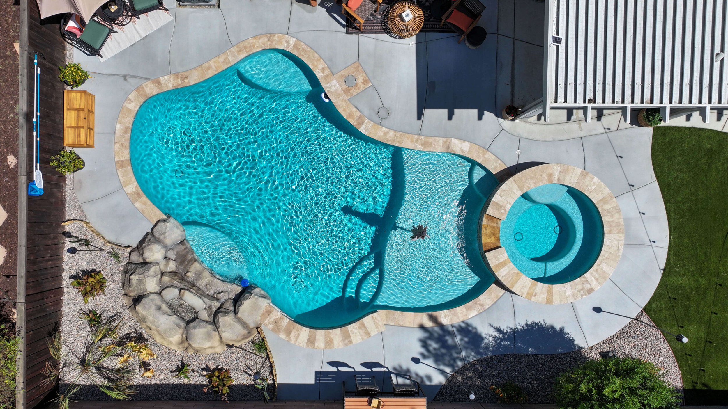 An aerial view of a backyard swimming pool with a water slide, surrounded by a concrete deck, lounge chairs, a wooden bench, and a small landscaped garden area with plants and rocks.