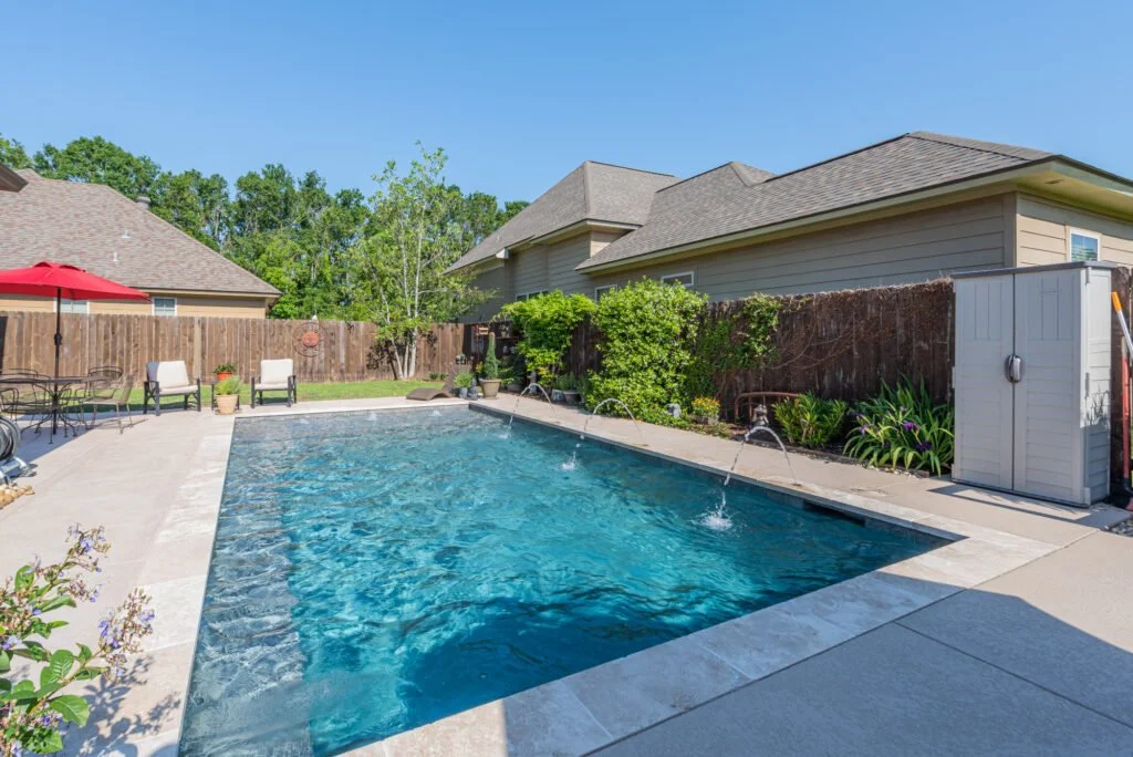 Backyard with a rectangular swimming pool, outdoor furniture, a red umbrella, a wooden fence, and a shed under a clear blue sky.