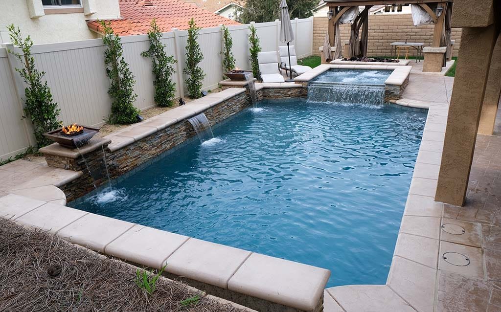 A backyard swimming pool with a built-in waterfall feature, surrounded by a stone and concrete deck, with a white privacy fence and greenery along the fence line.