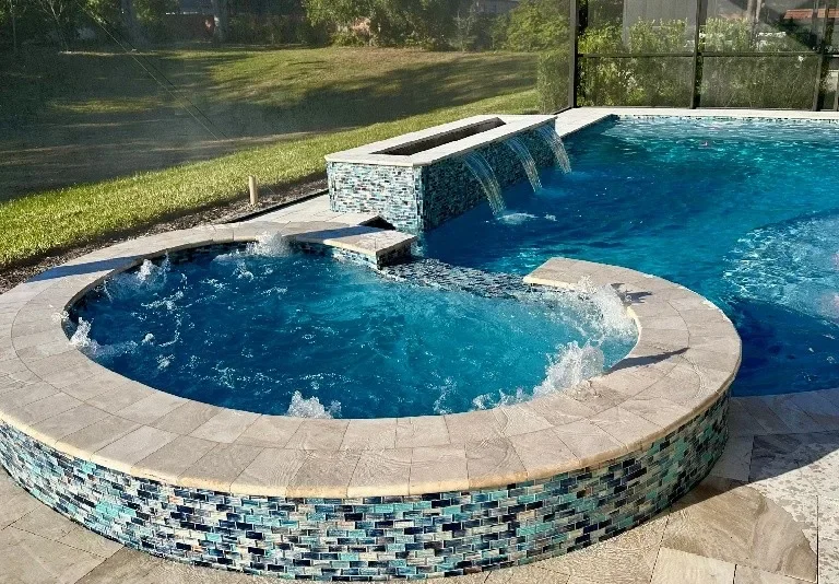 A backyard swimming pool with a built-in hot tub, featuring a waterfall feature and a tiled exterior, surrounded by a patio and a grassy lawn.