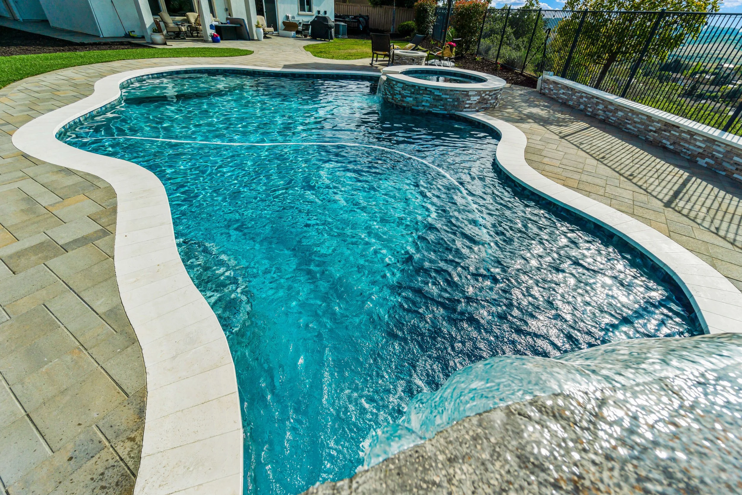 A backyard with a kidney-shaped swimming pool, a hot tub, patio furniture, and a fence, with trees and a neighboring house visible in the background.