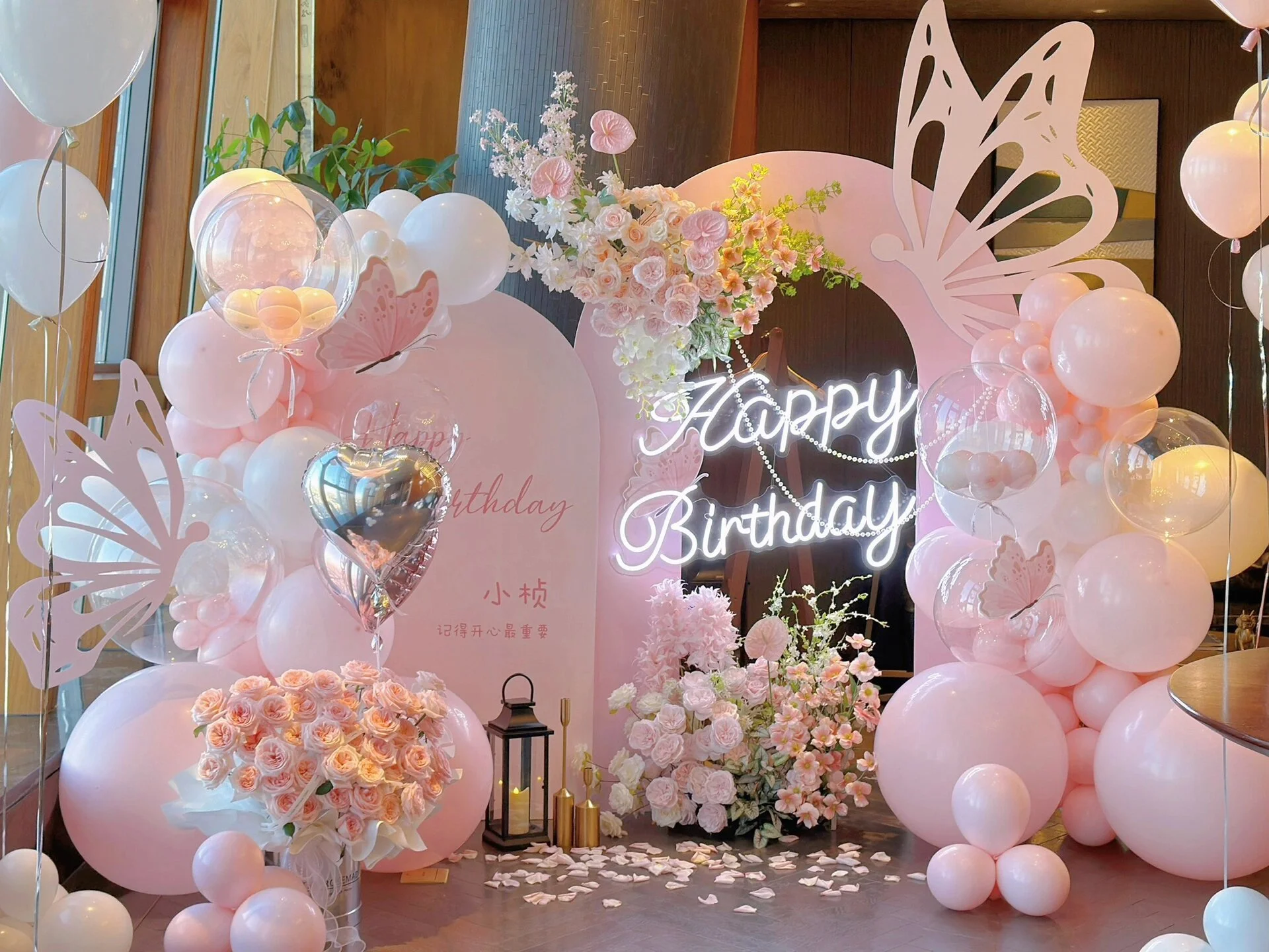 Decorative pink and white birthday celebration setup with balloons, pink roses, butterfly cutouts, a neon sign that says 'Happy Birthday,' and a floral arrangement.