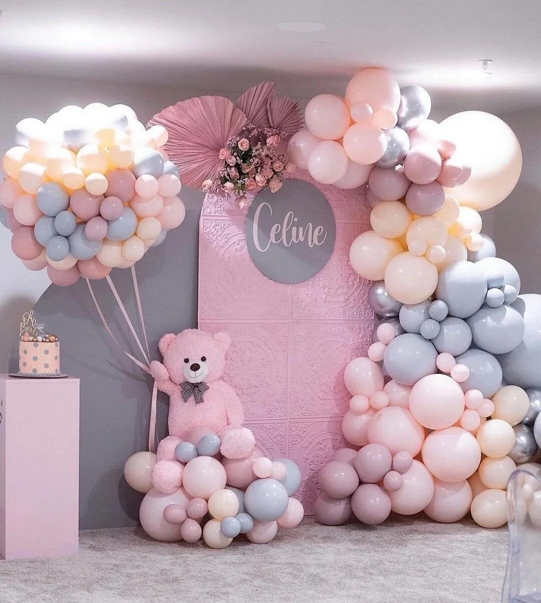 Decorative backdrop for a celebration featuring pastel pink, blue, white, and silver balloons, with a pink teddy bear, pink flowers, and a pink sign with the name 'Celine' in white cursive. There is a small pink and white polka dot cake on a pink ped