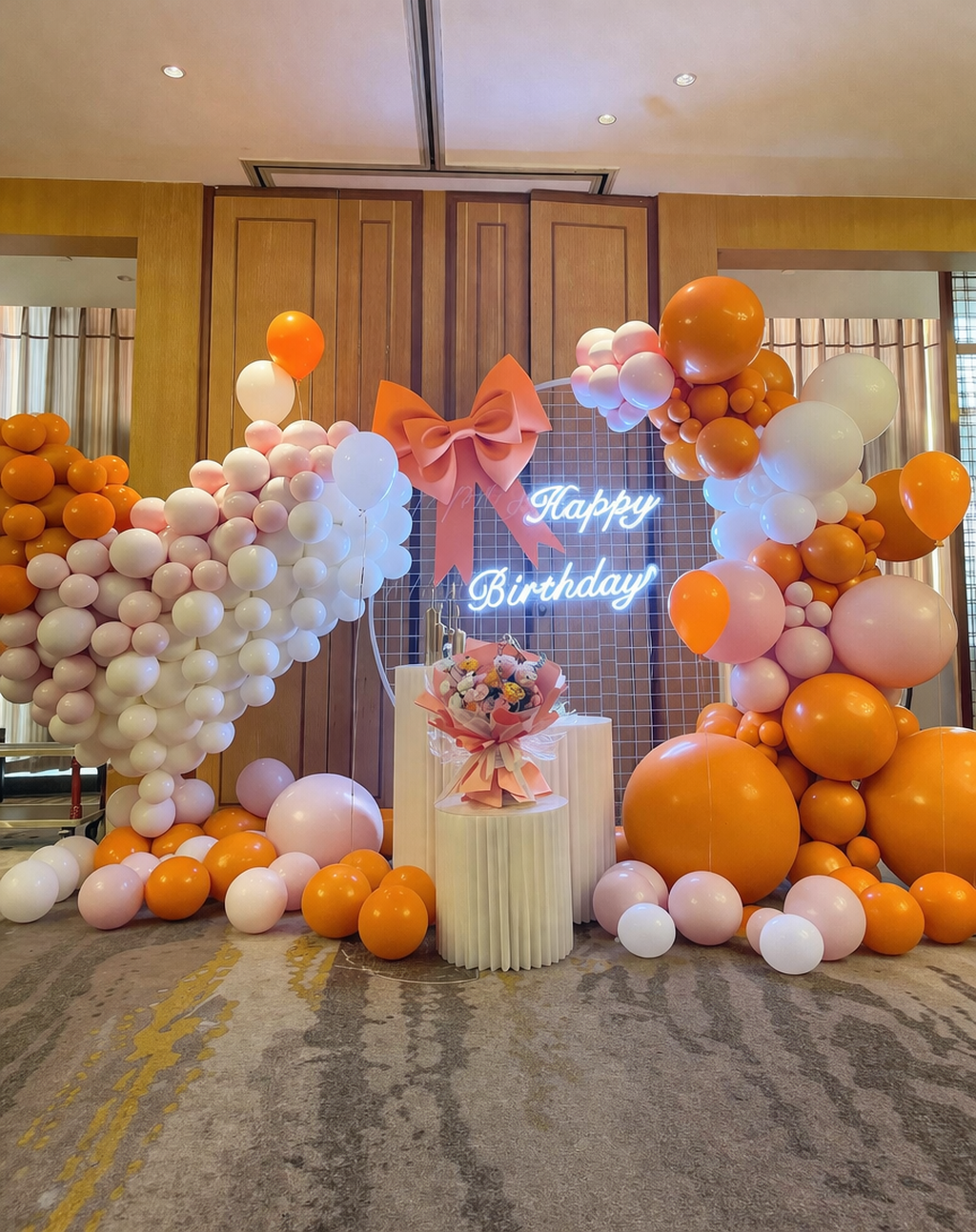 Decorative birthday display with orange, pink, and white balloons arranged on either side of a small table with a cake, backdrop featuring a neon "Happy Birthday" sign, and a large pink bow at the top center.