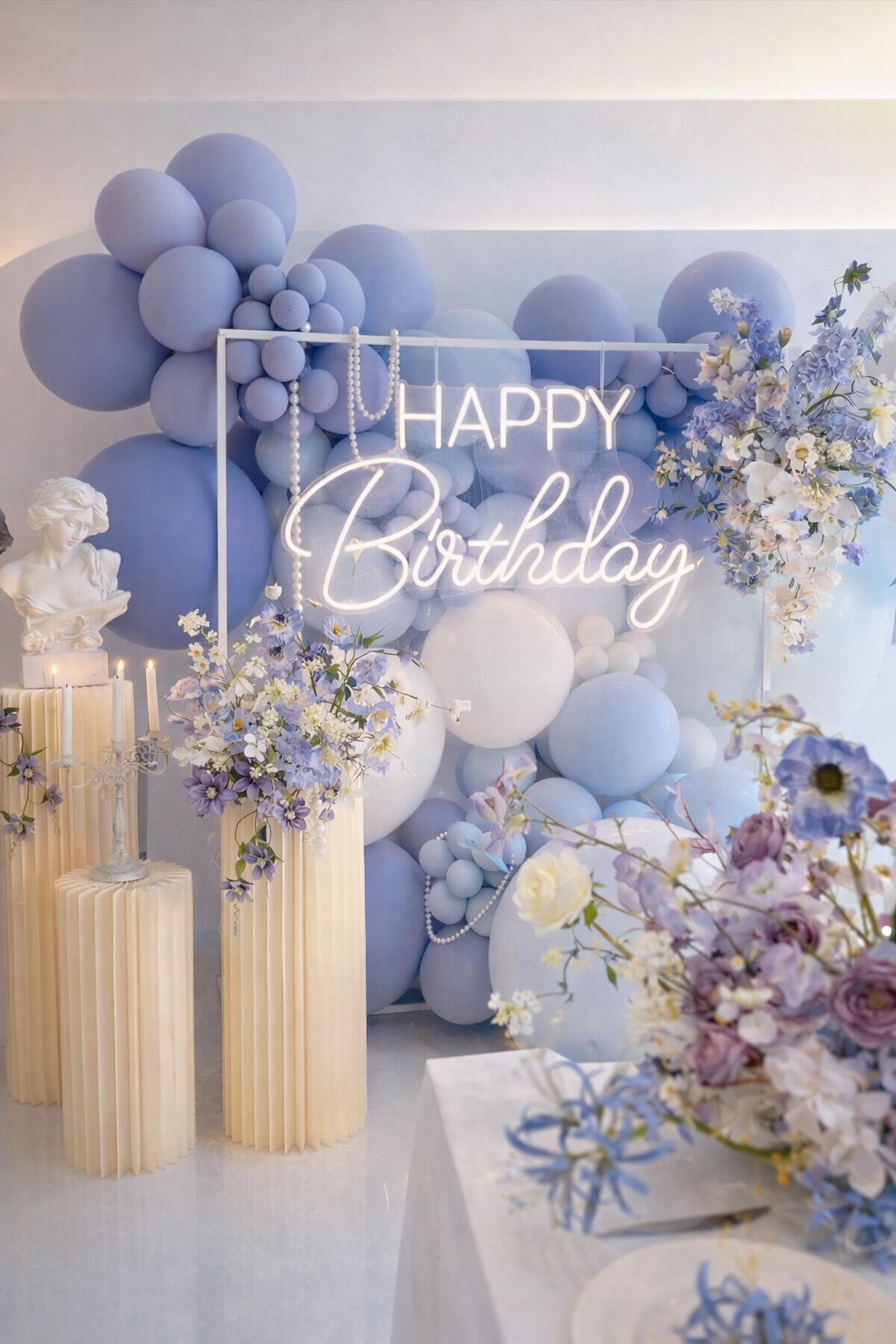 Blue and white balloon backdrop with a neon "Happy Birthday" sign, surrounded by floral arrangements and white decorative statues.