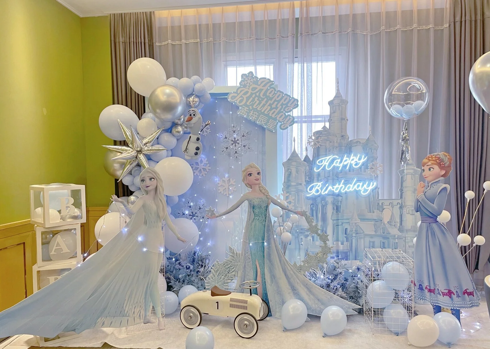 Frozen themed birthday party setup with characters Elsa, Anna, and Olaf, white and blue balloons, snowflake decorations, and a "Happy Birthday" sign.