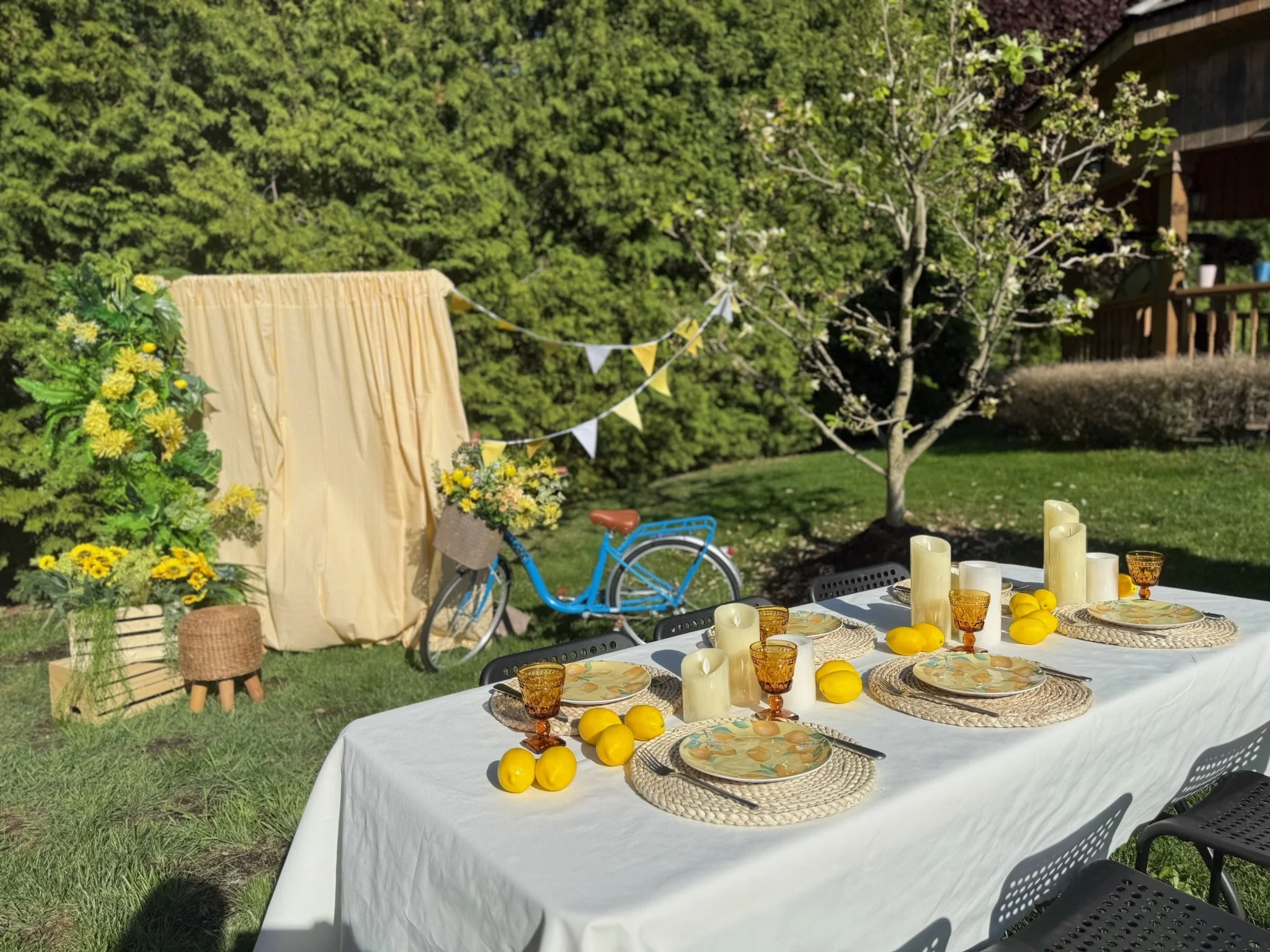 A decorated outdoor garden party setup with a long table covered in a white tablecloth, set with plates, glasses, and candles; yellow lemons are used as decorations. In the background, a blue bicycle with a flower basket, a beige fabric backdrop, and