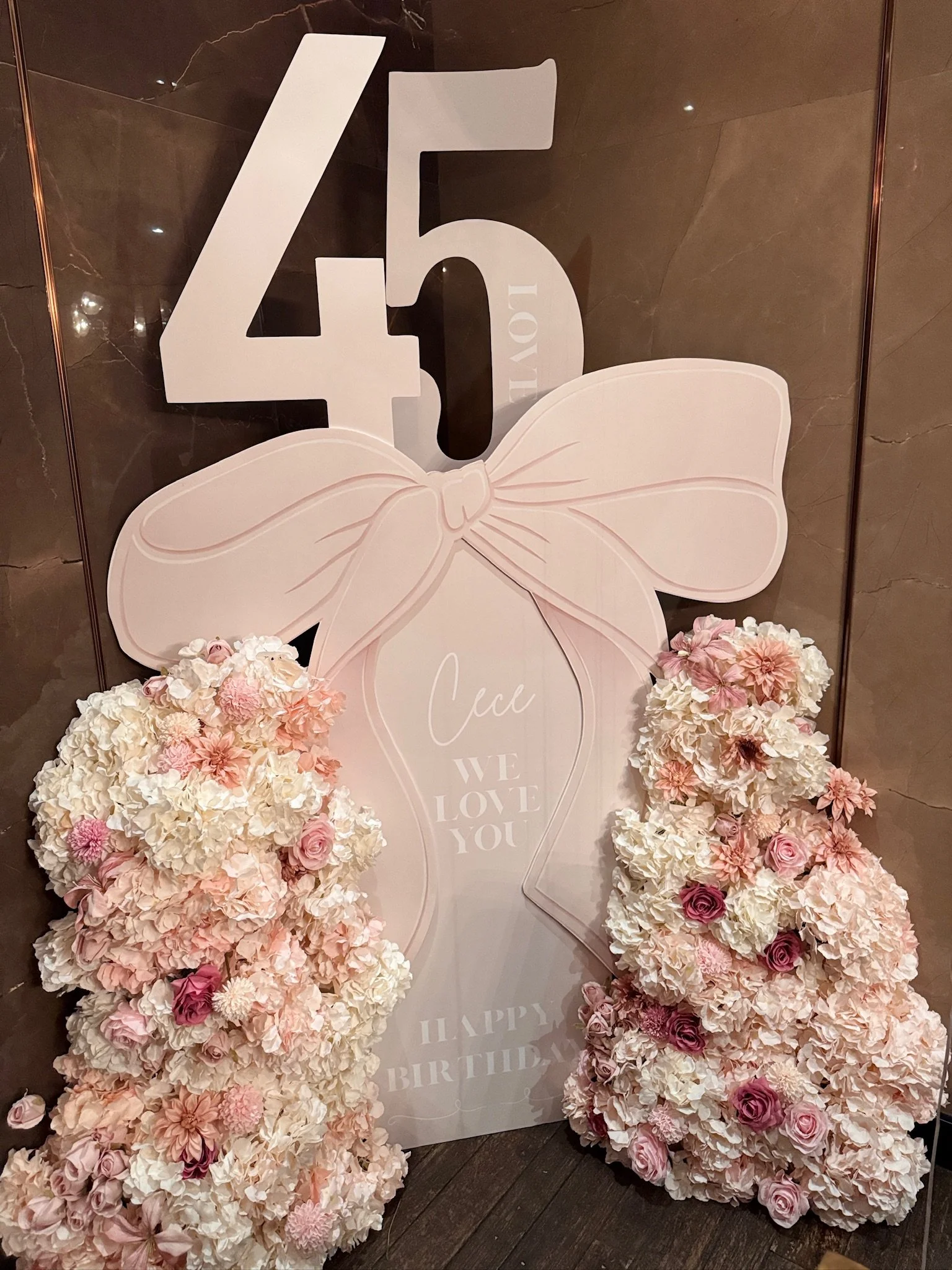 Pink and white birthday decor with large number 45, floral arrangements of pink and white flowers, and a sign with the words 'Coco, We Love You' and 'Happy Birthday' behind a large pink bow.