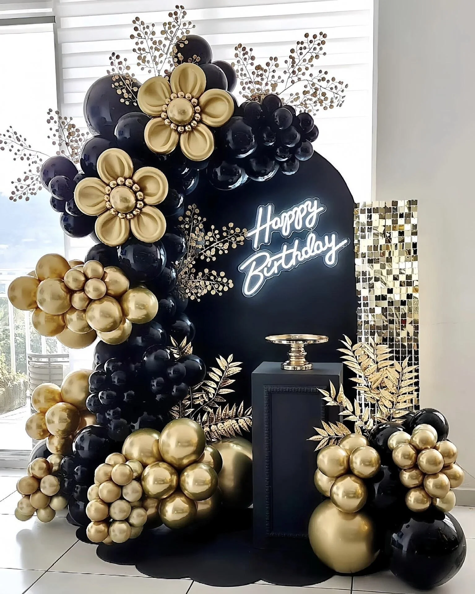Black and gold balloon arch with gold flower and leaf decorations, illuminated 