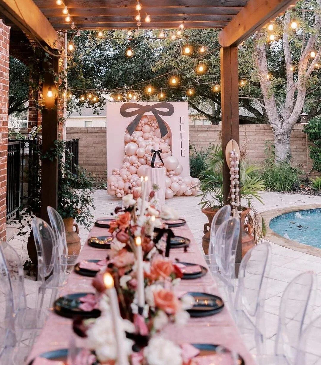 Outdoor birthday party setup with a long table decorated with candles and flowers, string lights overhead, a backdrop with pink and white balloons, a large gift box prop, and a swimming pool nearby.