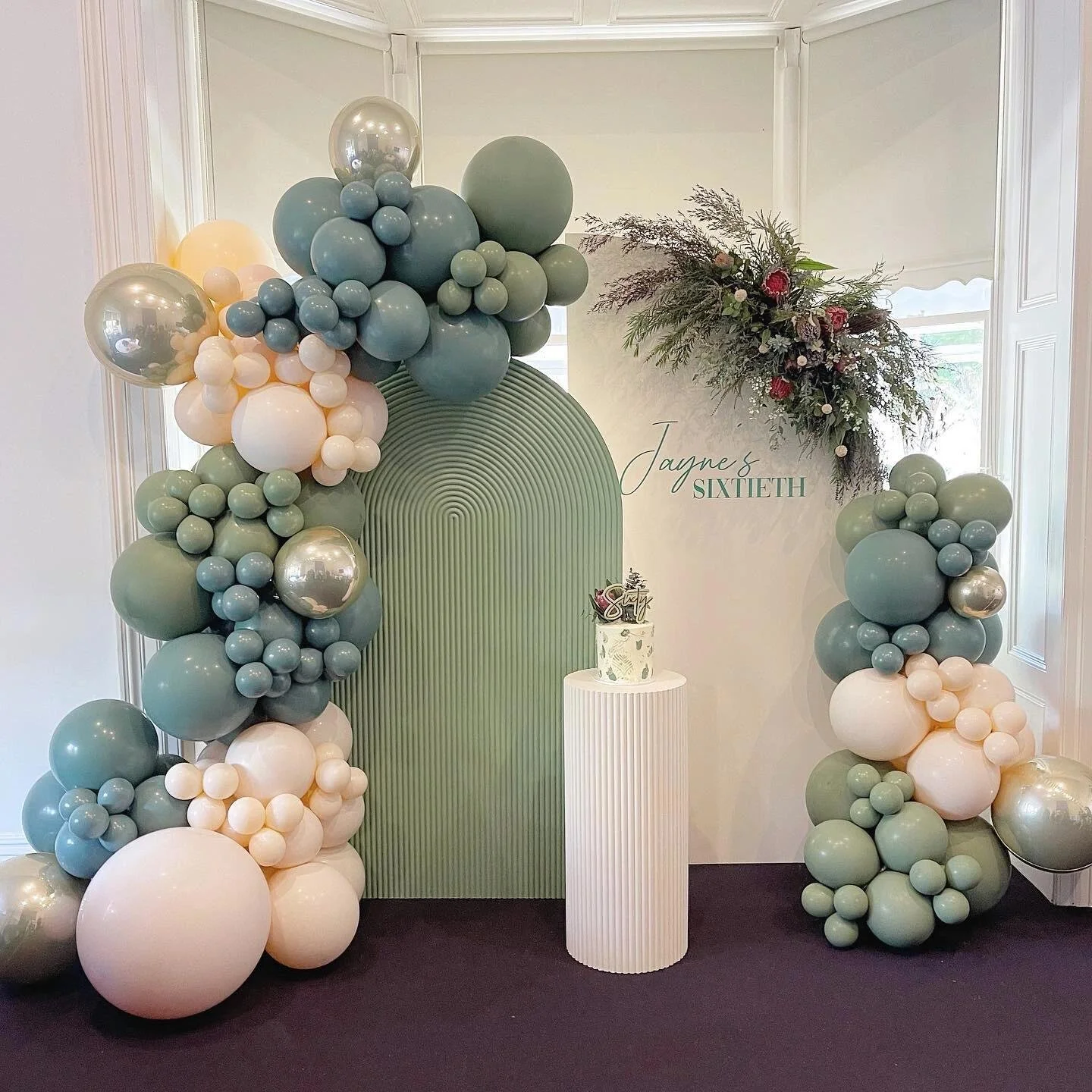 Birthday celebration setup with a green balloon arch, floral arrangement, cake on a white pedestal, and a sign that says "Jayne's Sixtieth" in a decorated room.
