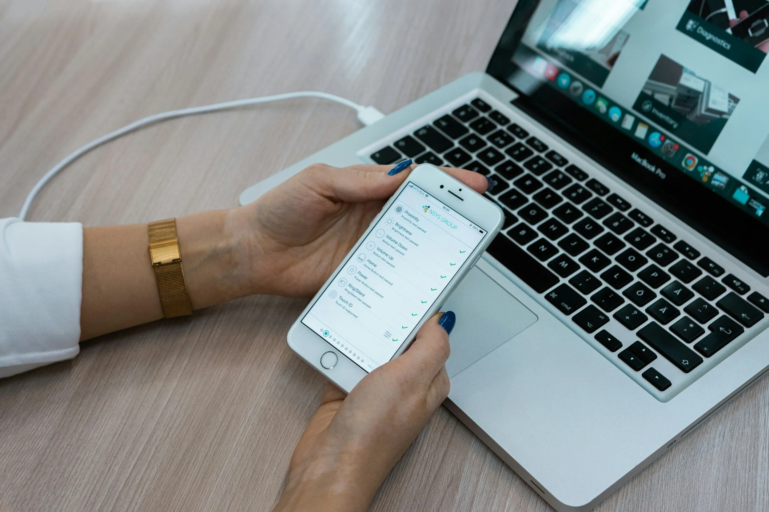 A person holding a smartphone with a hands-free device settings screen open, beside a laptop on a wooden table.