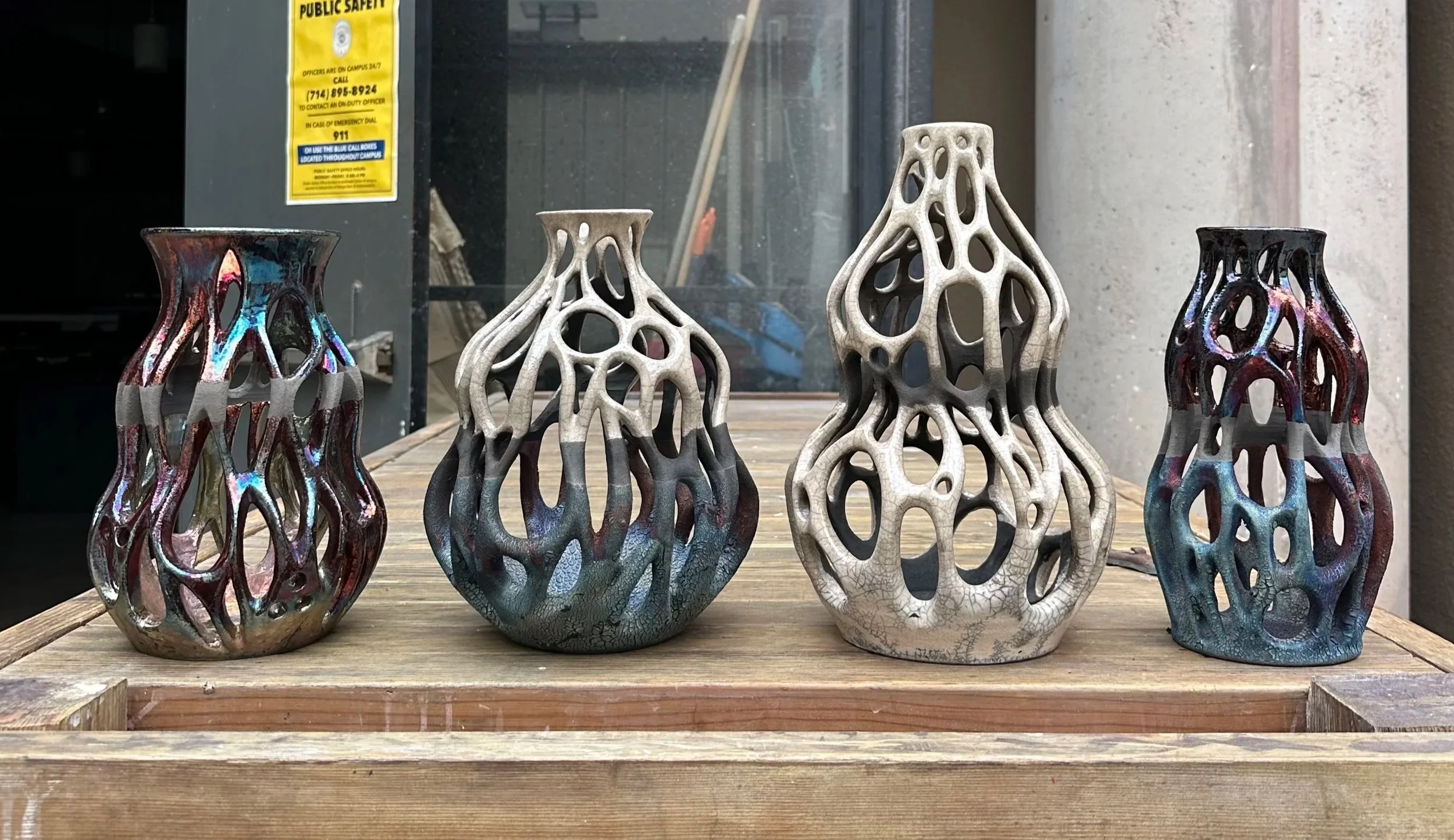 Four decorative ceramic vases with organic, hollow, web-like patterns, two with iridescent finishes and two with matte finishes, displayed on a wooden table.