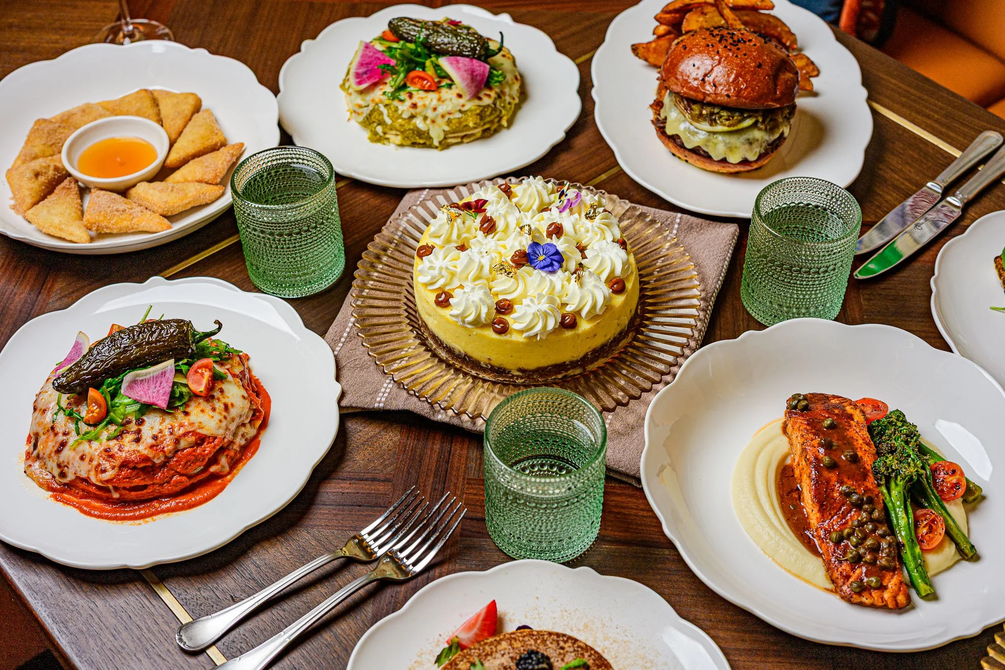 A table set with various dishes including a cheese cake, a burger, fried appetizers with dipping sauce, and plates with pasta and chicken, along with glasses and utensils.